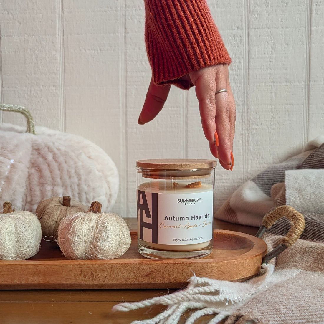 A fall scented candle, Autumn Hayride, on a table with a fall blanket and a couple fall pumpkin decorations and a hand reaching down to the candle.