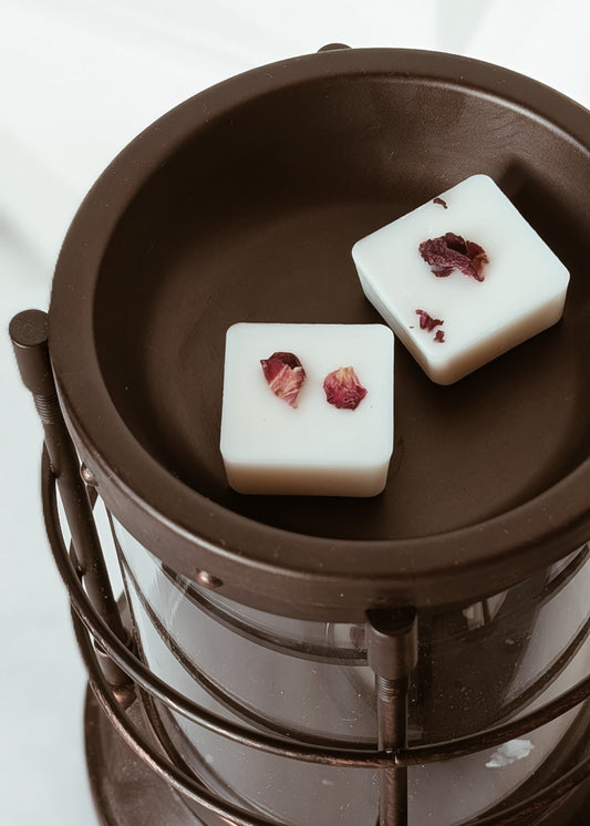 Two white square wax melts with red rose pedals on a rustic wax warmer