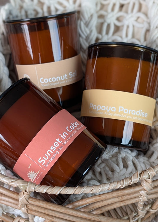 Three summer scented amber glass candles with labels in a woven basket on a textured surface