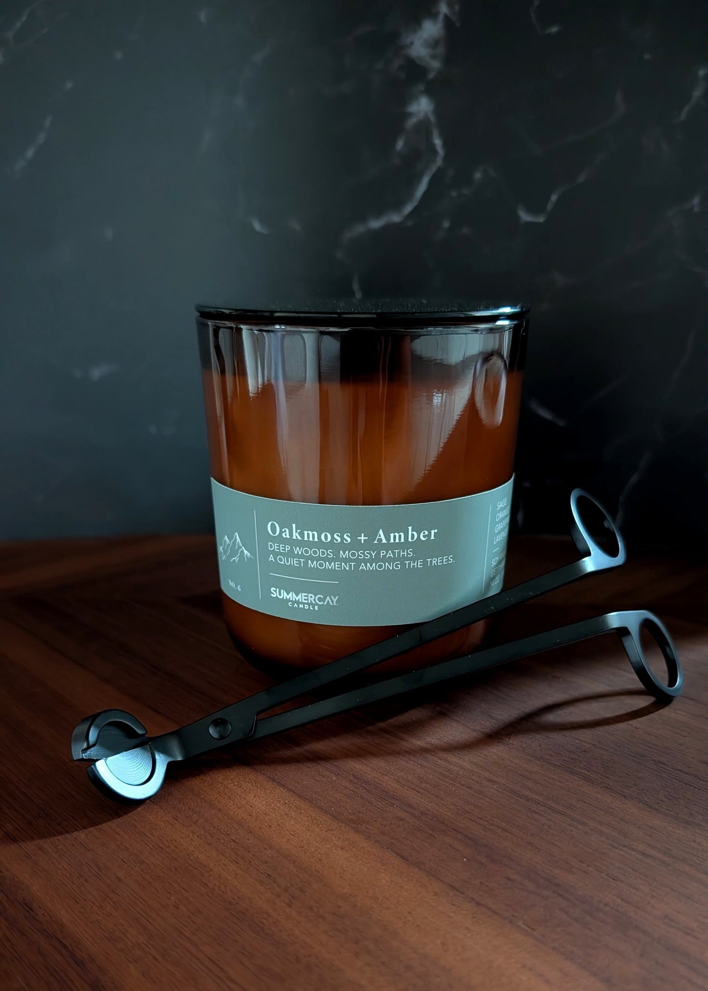 Candle labeled 'Oakmoss & Amber' on a wooden surface with a set of black wick trimmers with a dark background