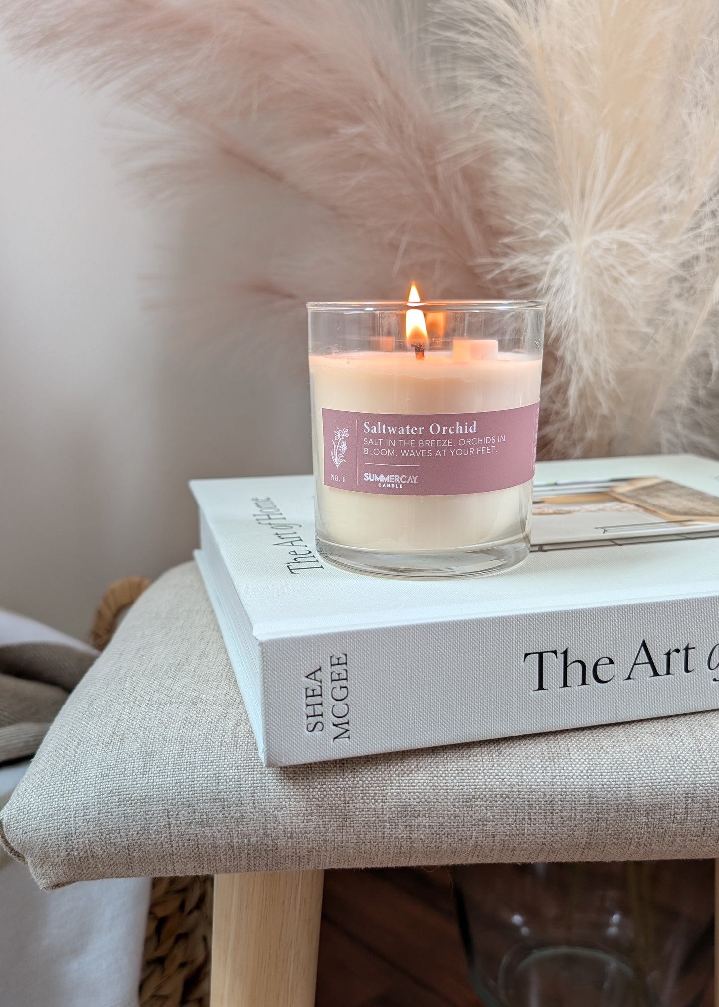 A lit candle in a clear glass jar sits on top of a book on a beige upholstered stool. The candle label reads 'Saltwater Orchid'.
