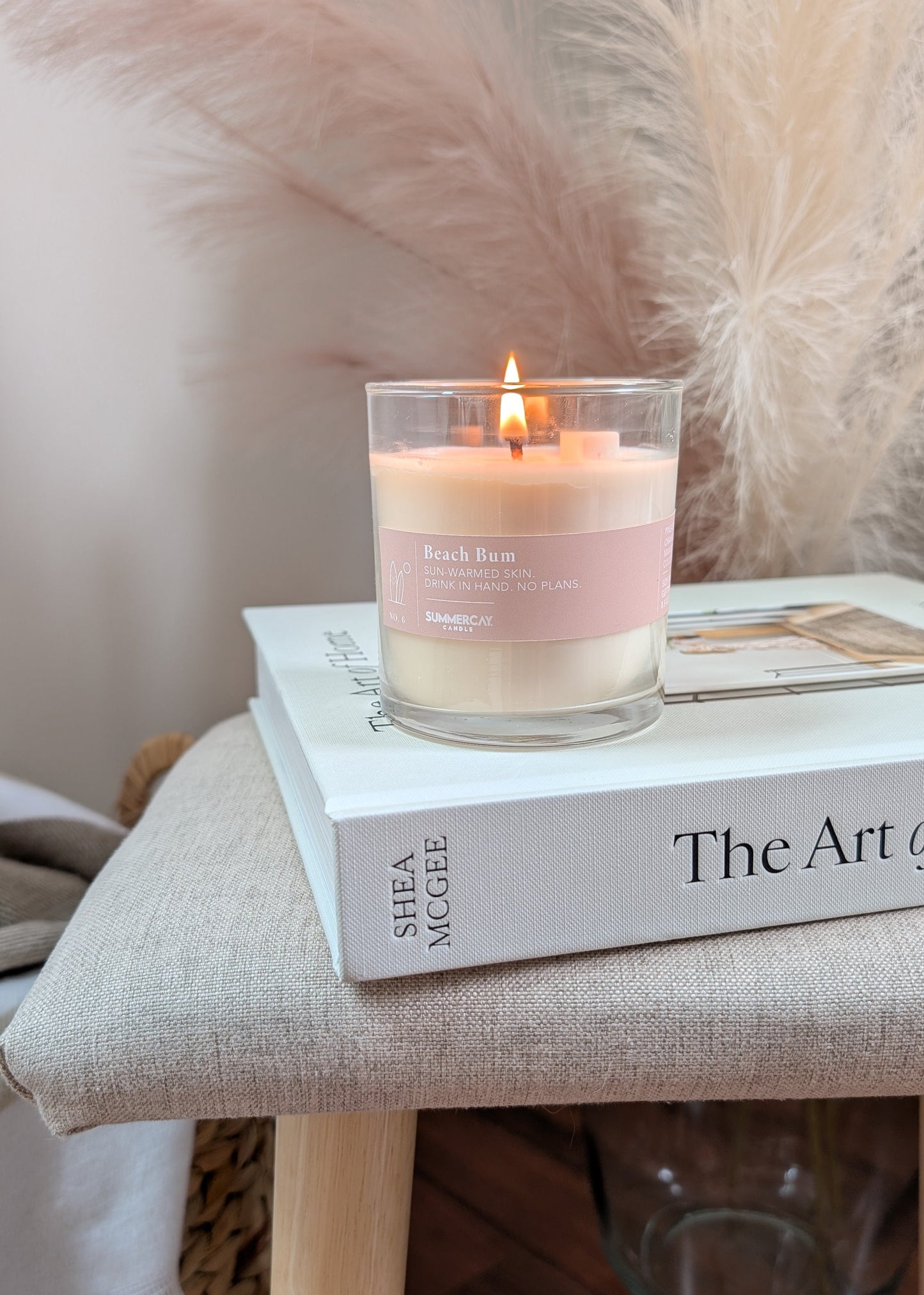 A lit candle in a clear glass jar sits on top of a white book on a beige upholstered stool, with pampas grass and a glass vase in the background.