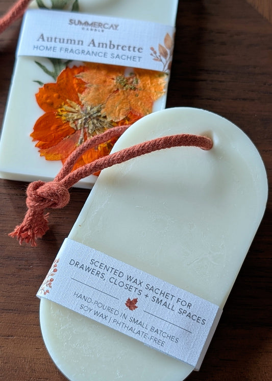An oval-shaped cream-colored home fragrance sachet wrapped in a white paper band, displayed on a dark wooden surface next to its matching printed packaging box.