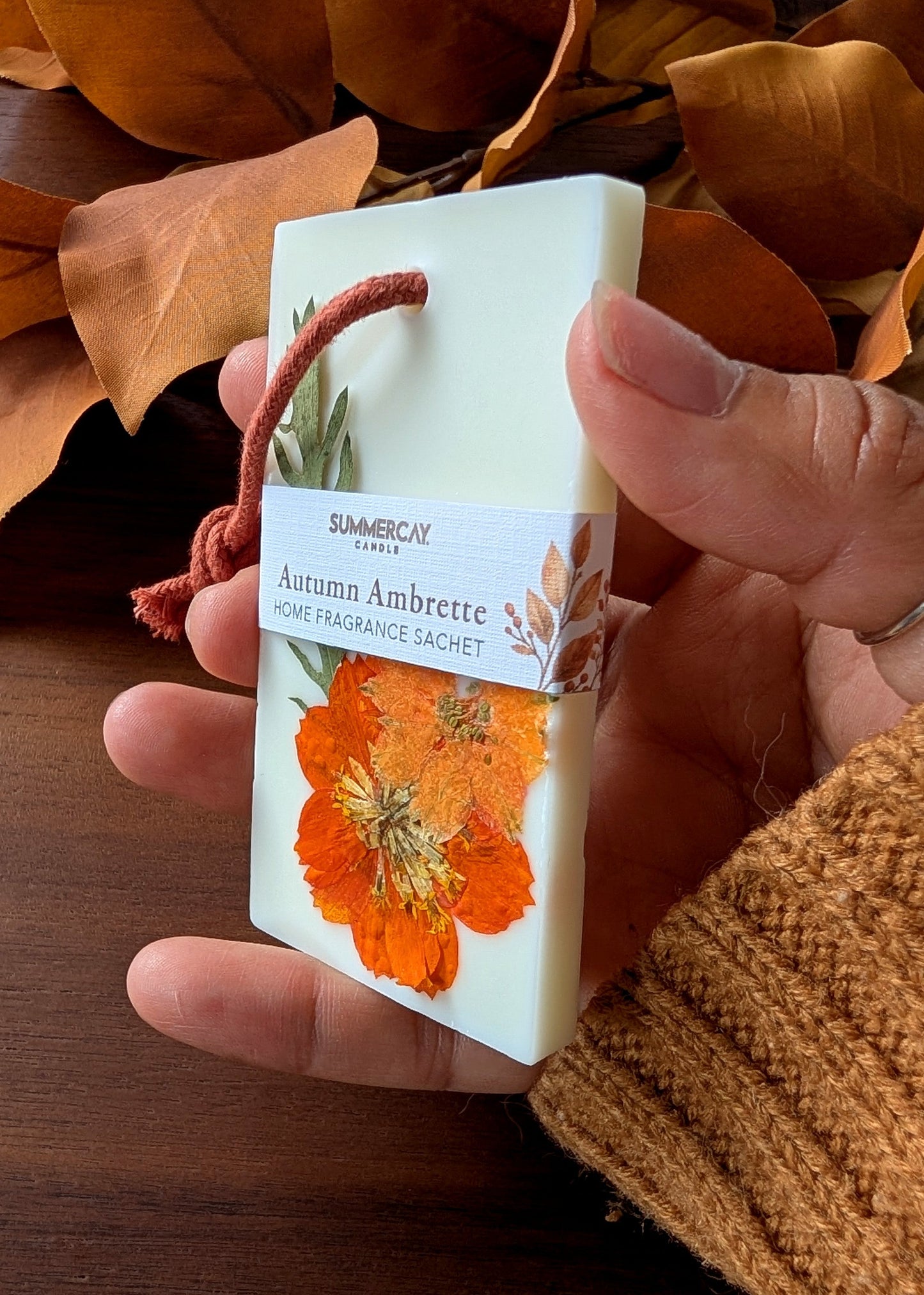 A hand holding a SummerCay Home Fragrance Sachet in Autumn Ambrette scent. The white rectangular sachet features a red hanging string, a white paper label with an orange flower, and is set against a background of dry brown autumn leaves and a wooden table.