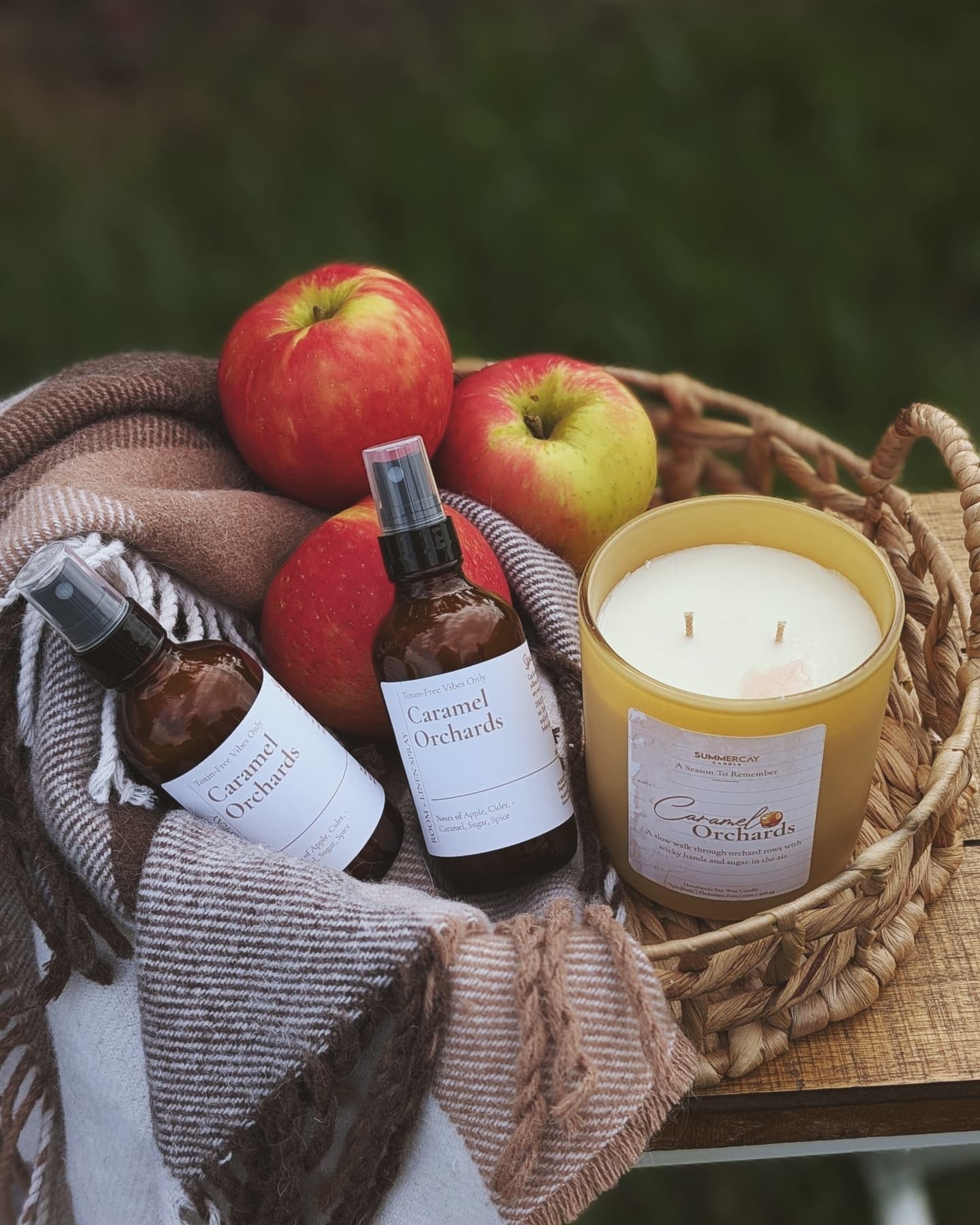 Caramel Orchards products including a candle, bottles, and apples on a woven basket with a blanket.