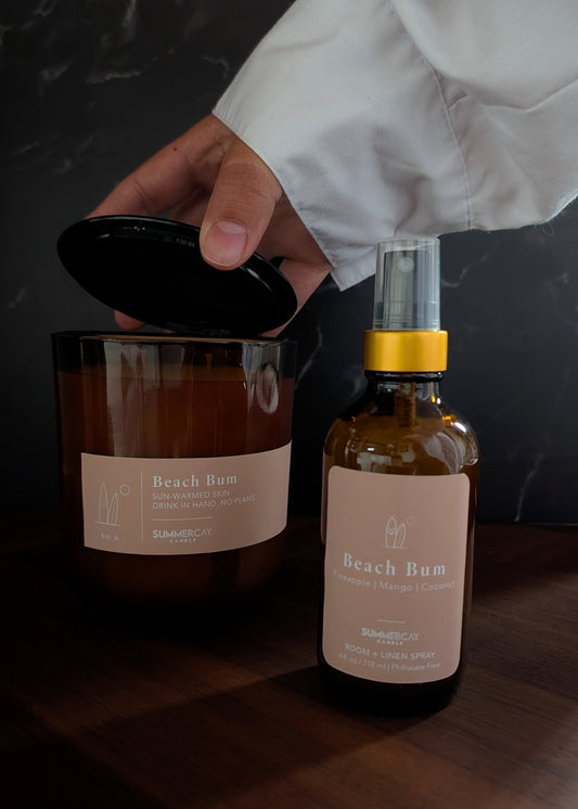 Beach bum candle and matching room + linen spray on a wooden table with a dark background and a hand pulling the candle lid off.