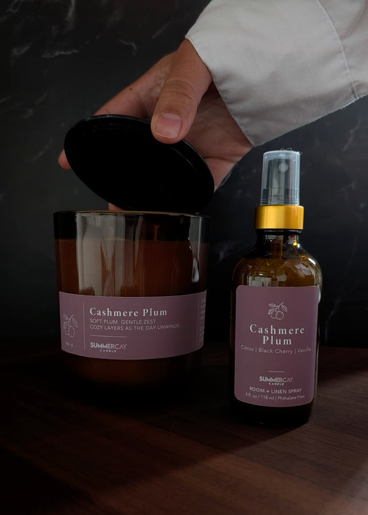 Cashmere plum amber candle with matching room + linen spray and a hand pulling of the candle black lid.