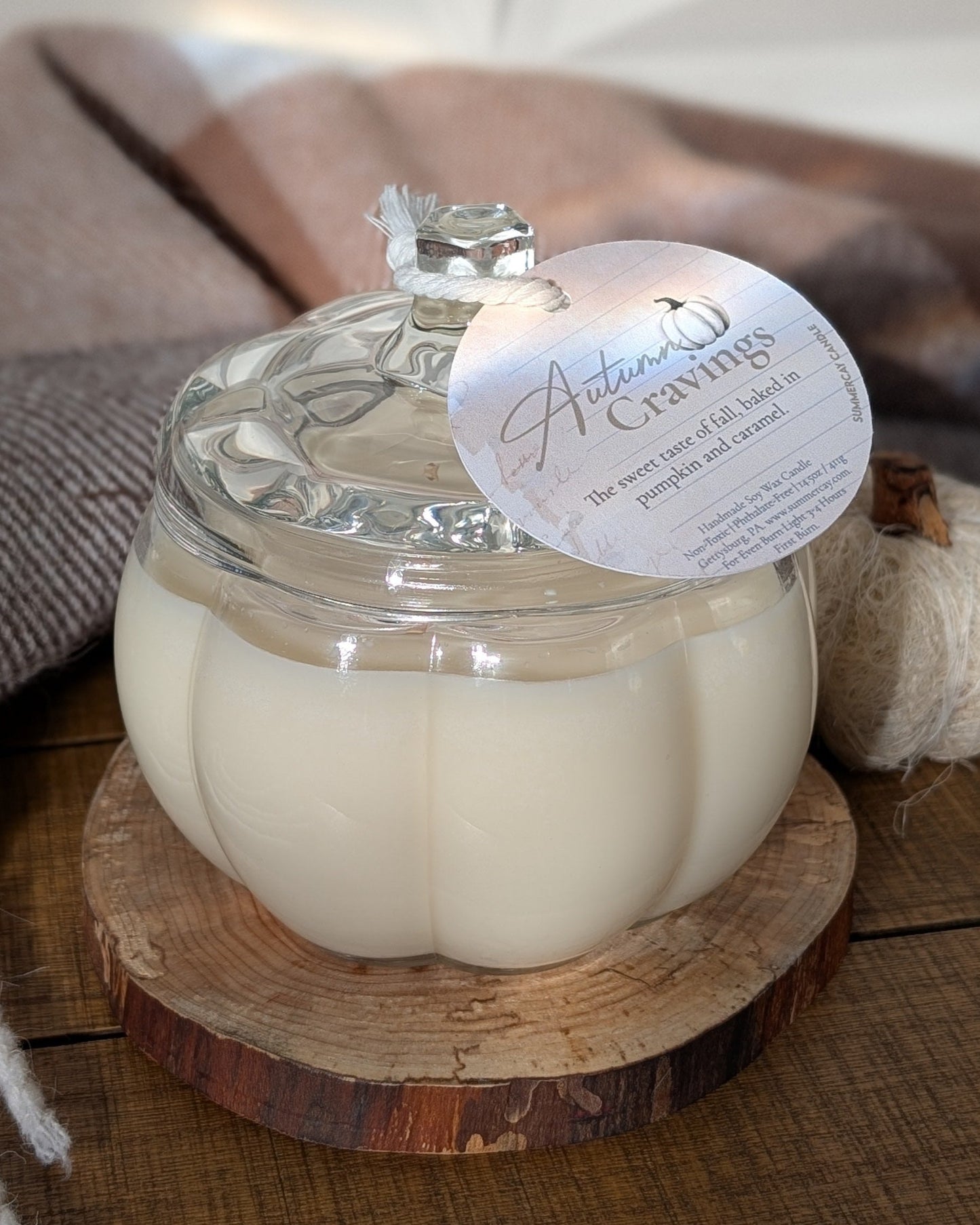 Glass jar candle on a wooden coaster with a tag, surrounded by a blanket and small pumpkin.