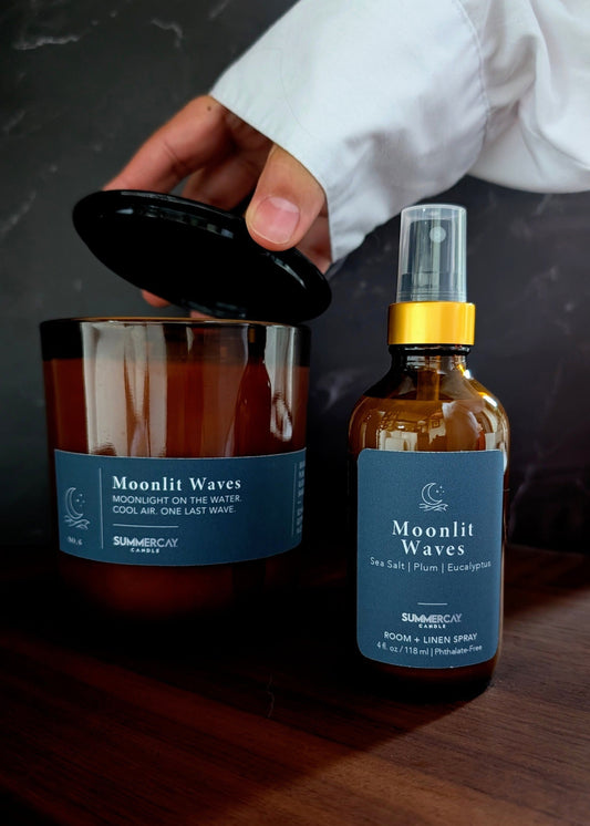 Moonlit Waves candle with matching room + room spray on a wooden table and a hand pulling the black candle lid of on a dark background.