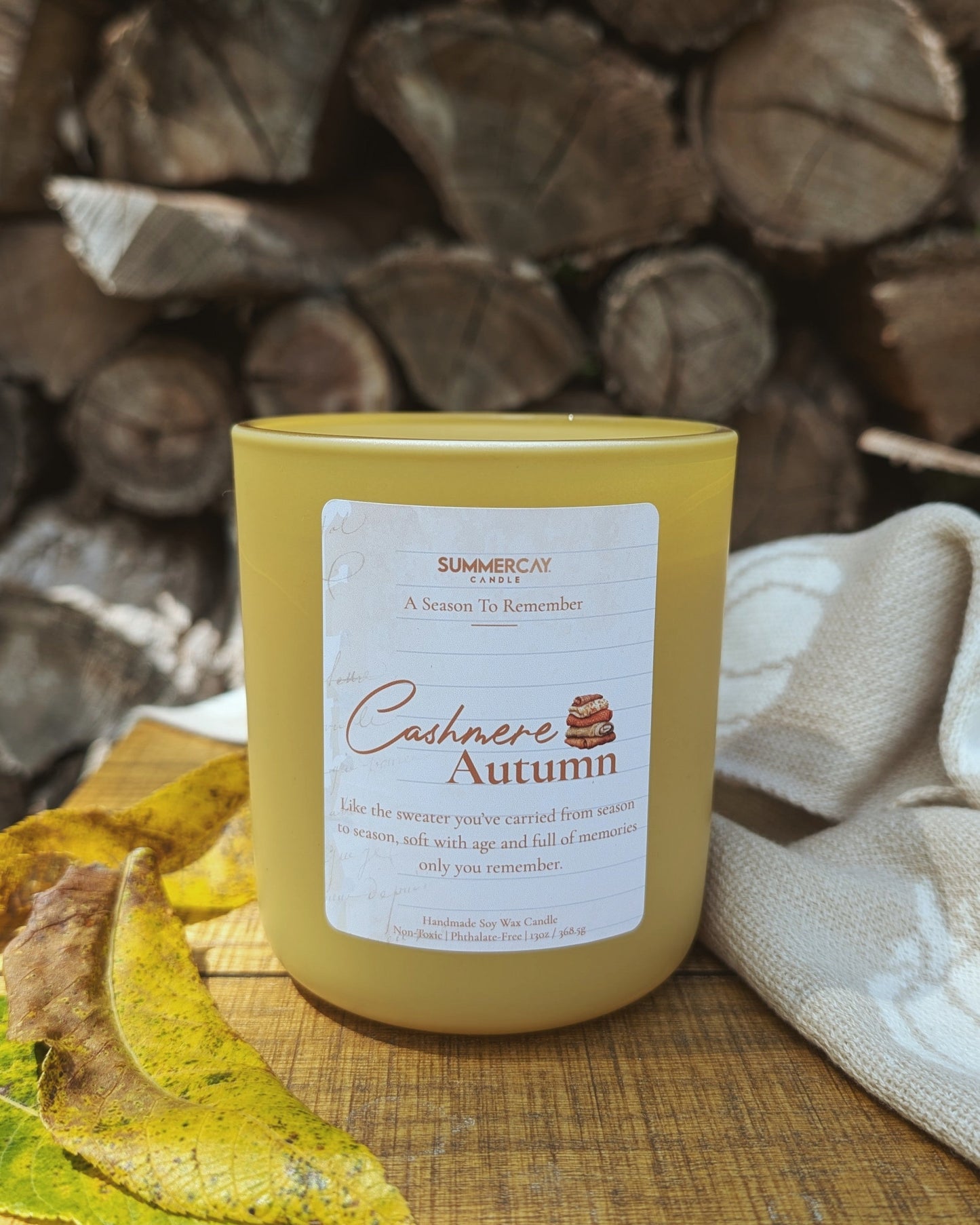 Yellow candle labeled 'Cashmere Autumn' on a wooden surface with logs in the background