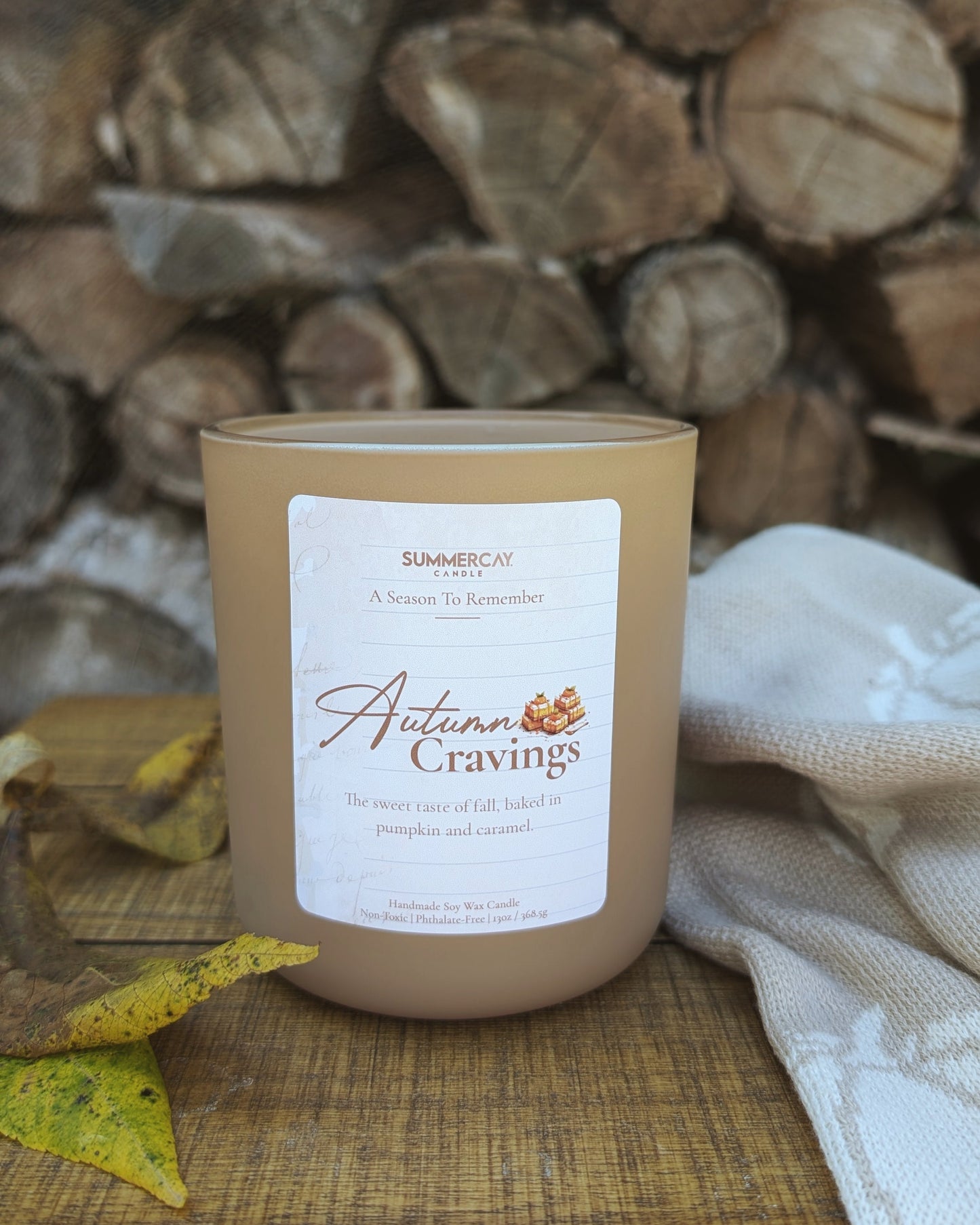 Candle labeled 'Autumn Cravings' on a wooden surface with stacked logs in the background