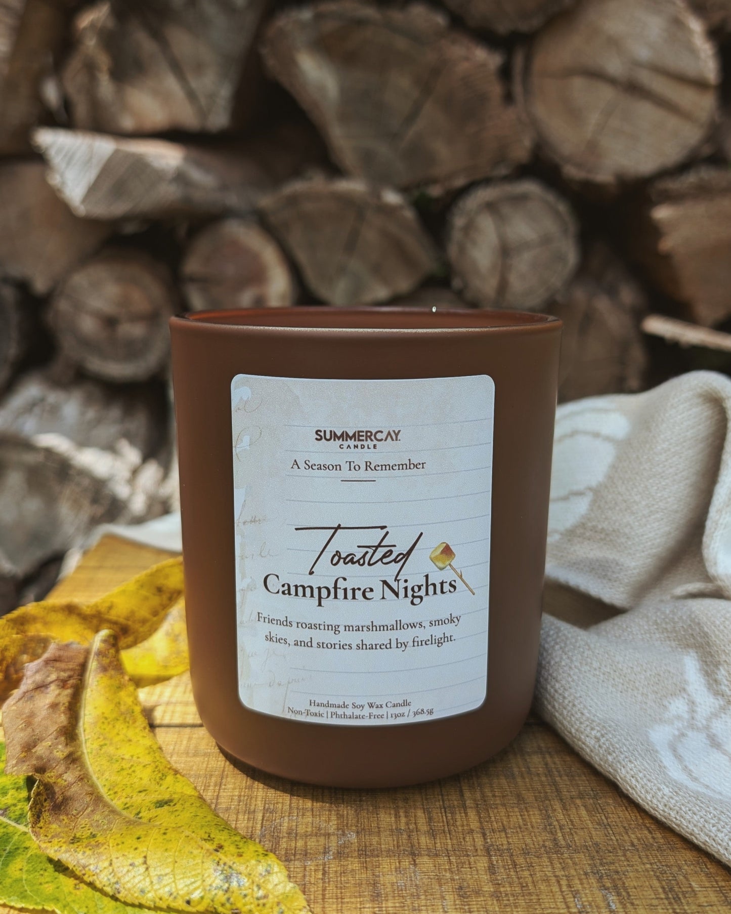 Candle labeled 'Toasted Campfire Nights' on a wooden surface with stacked logs in the background