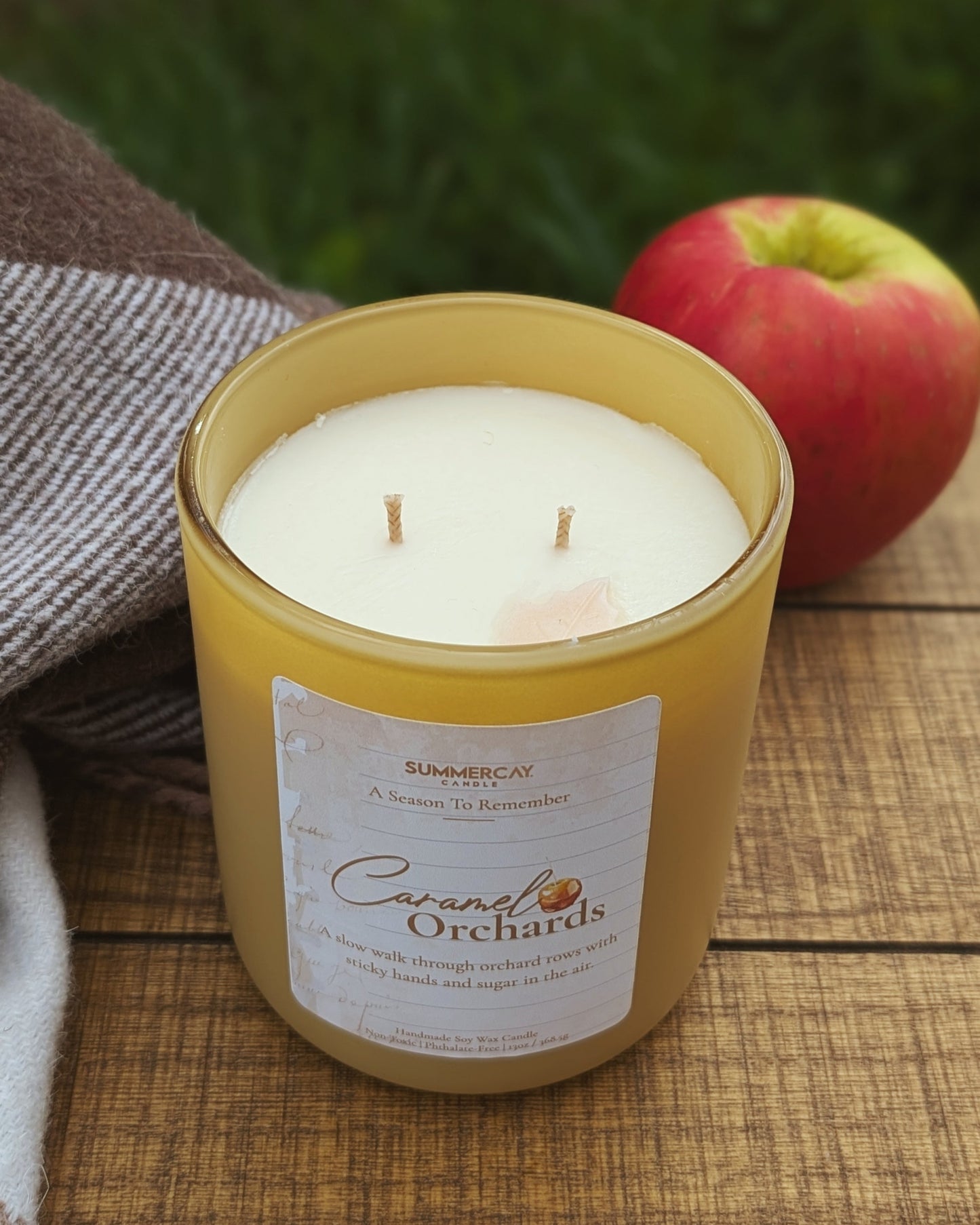 Candle in a yellow container with an apple and towel on a wooden surface
