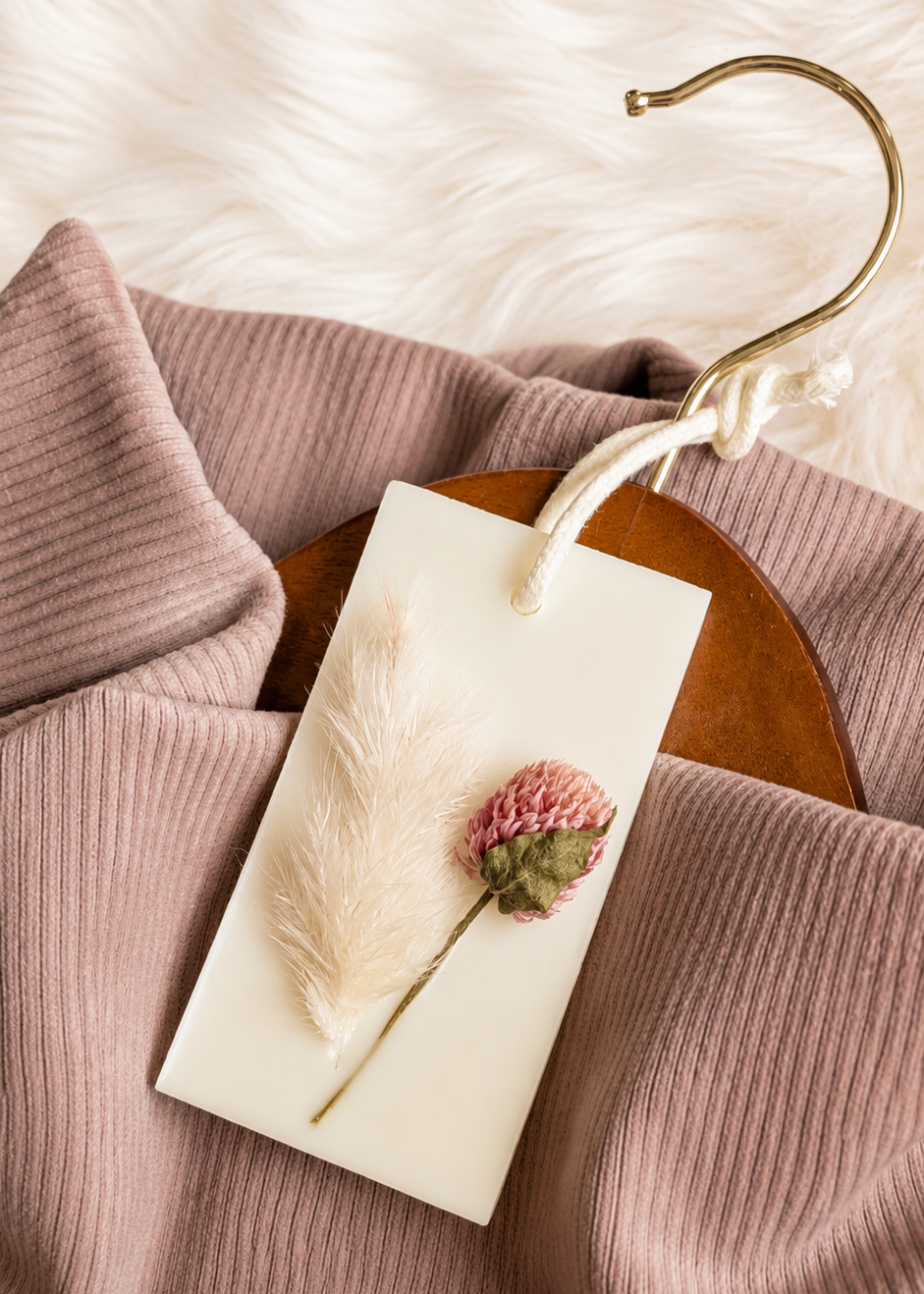 A dusty rose ribbed knit garment on a wooden hanger with a brass hook. A creamy white wax sachet with a dried pink flower and white feather.  Against a soft white faux fur background.
