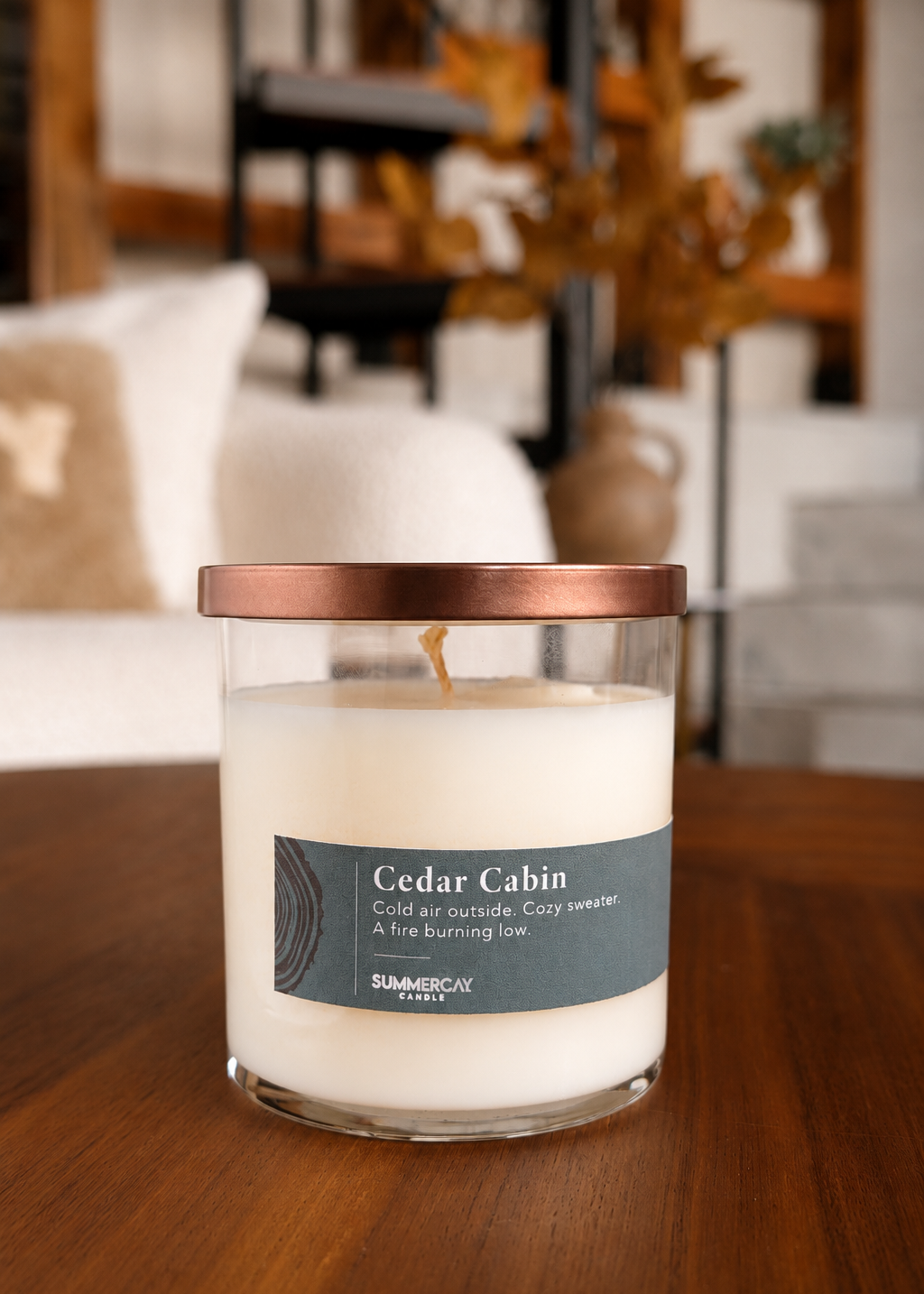 A Summerbly Candle in a clear glass jar with white wax and a bronze lid, labeled 'Cedar Cabin' sitting on a wooden table with a blurred living room background.