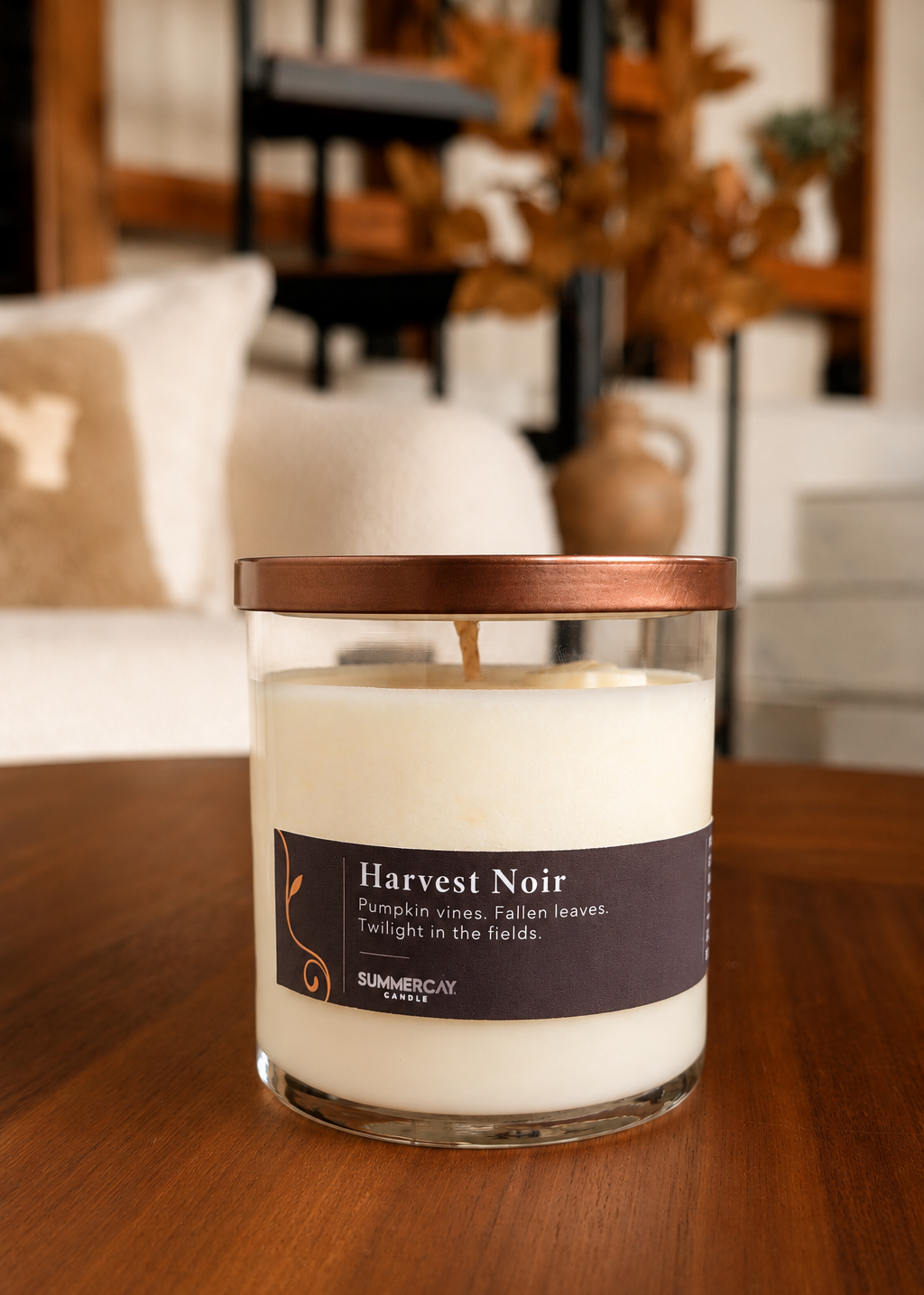 Harvest Noir scented candle in a clear glass vessel with a bronze lid sitting on a wooden table and blurred living room background.