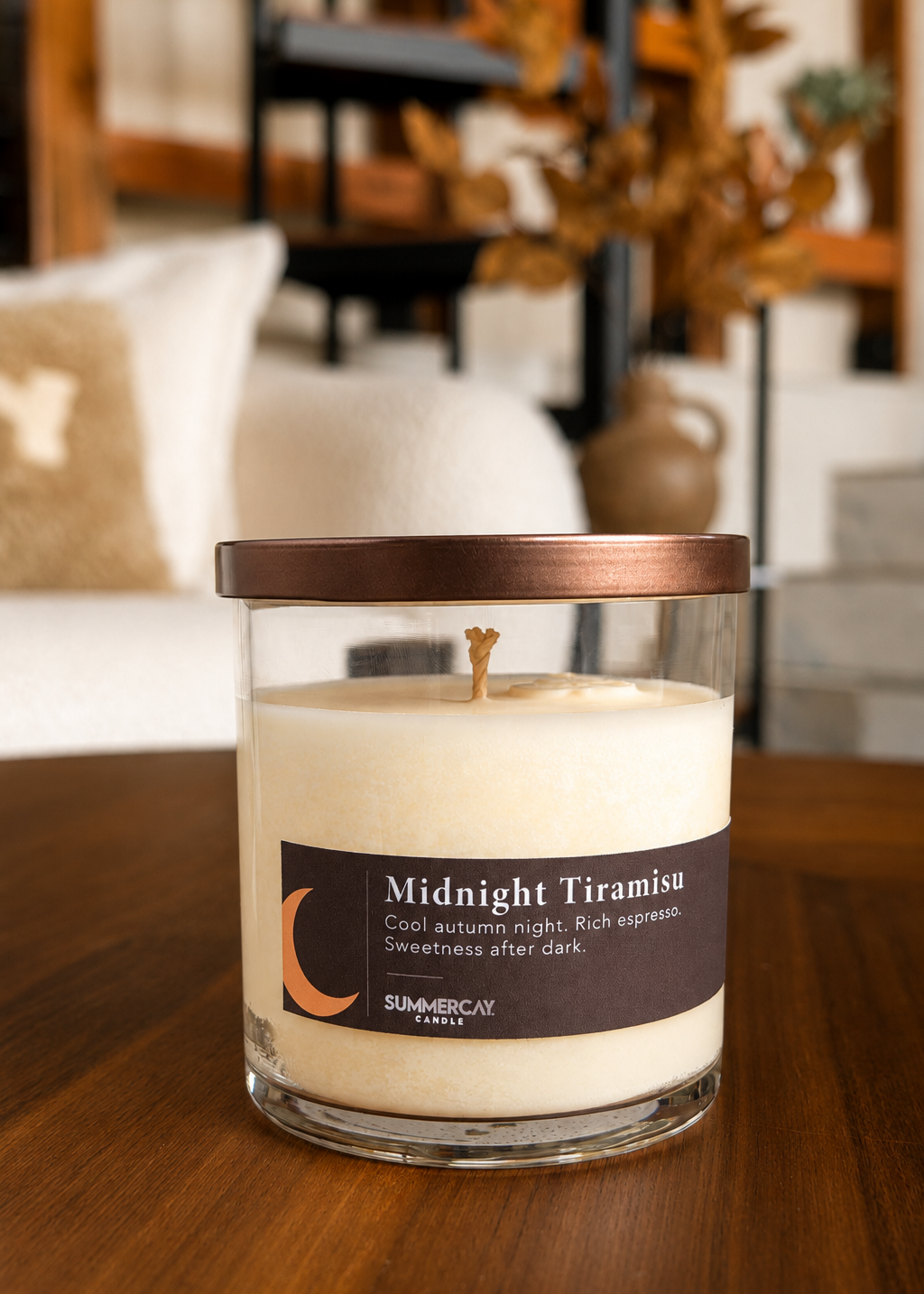 Mightnight Tiramisu soy candle in a clear vessel with a bronze lid. Sitting on a wood table with a blurred living room background.