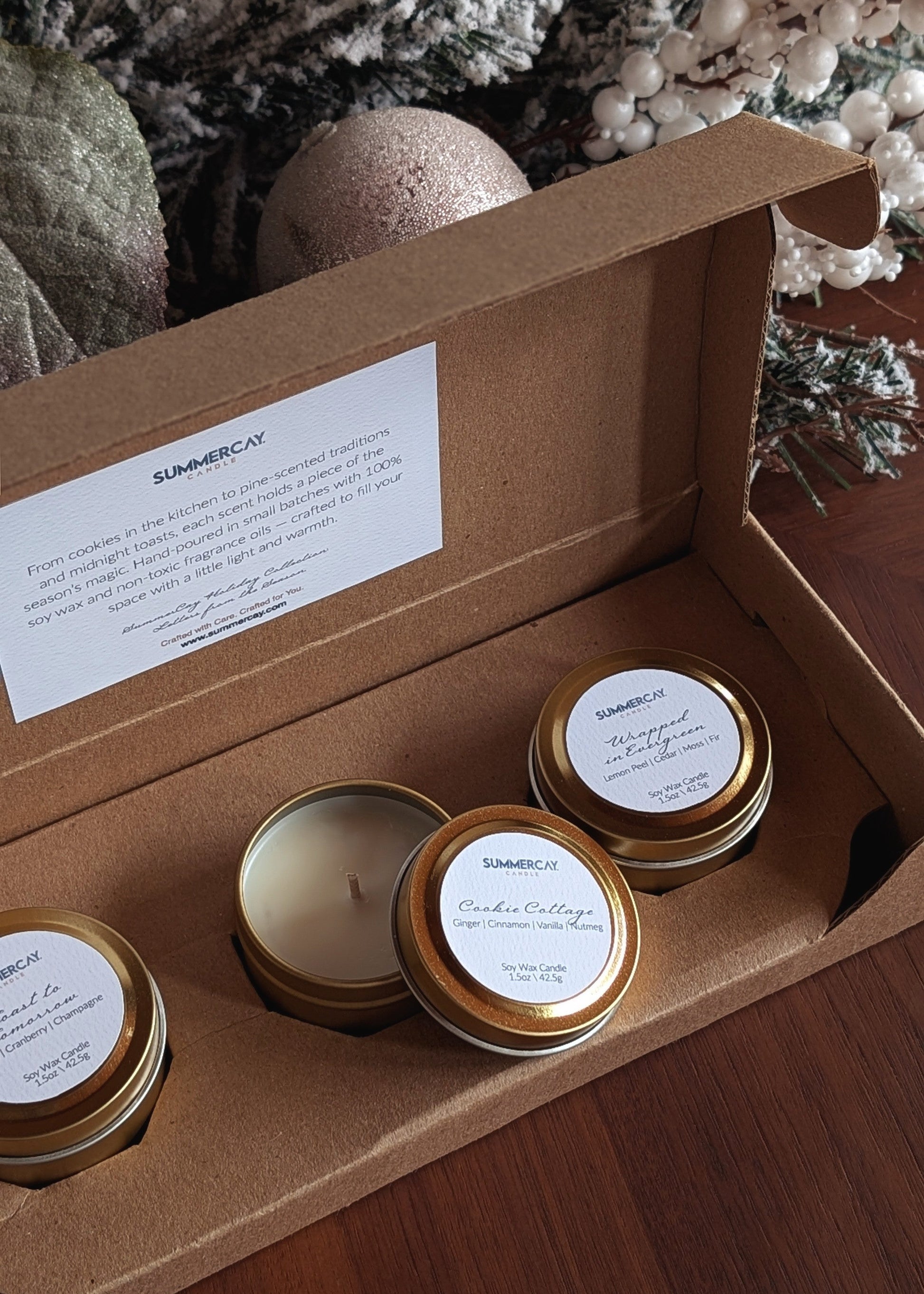 Four candles in gold jars with labels inside an open cardboard box, against a decorative background.