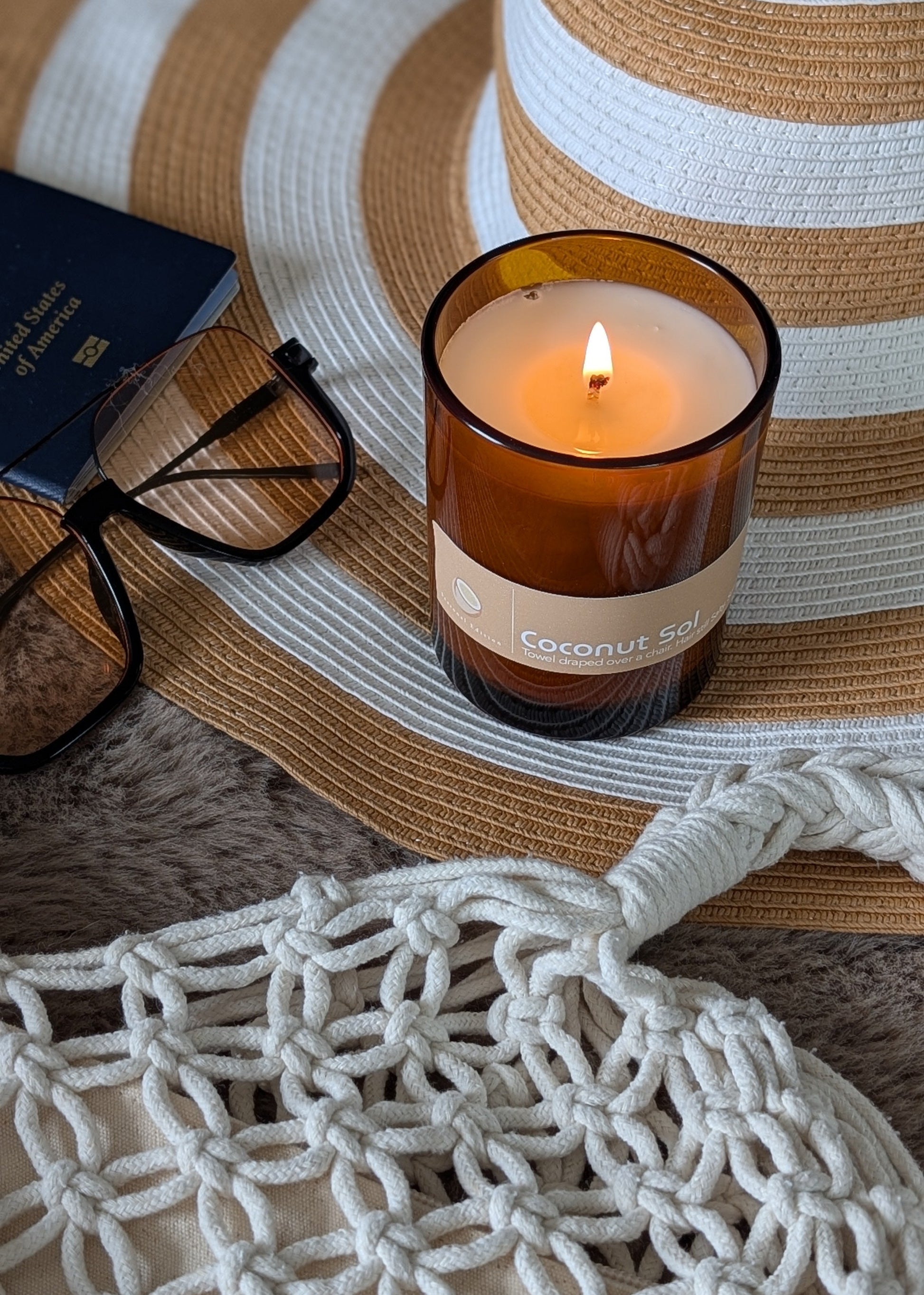 burning amber glass candle with beach items surrounding the candle. A summer hat, sunglasses, passport, woven bag.