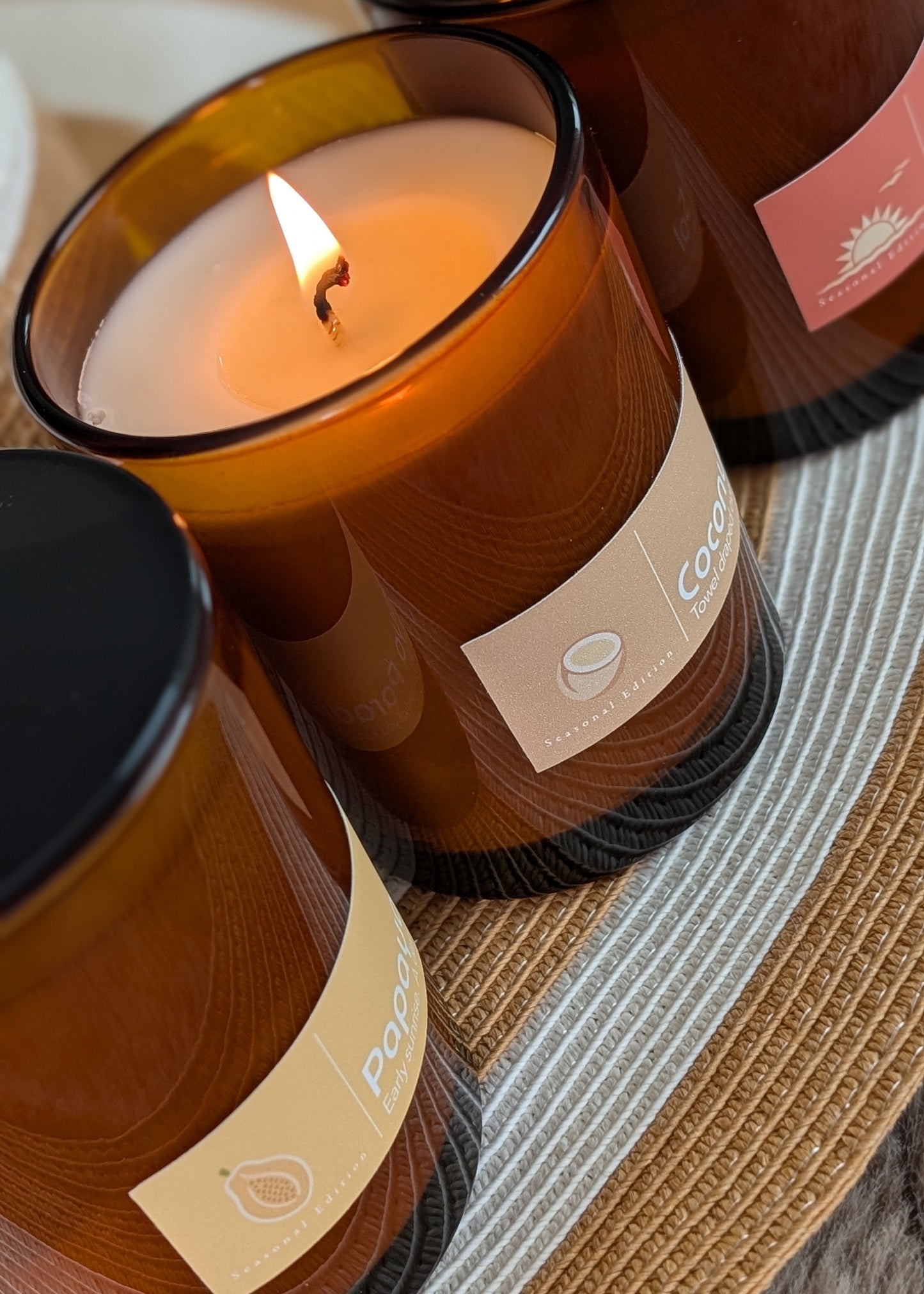 Three candles in brown glass jars with black lids on a woven mat.