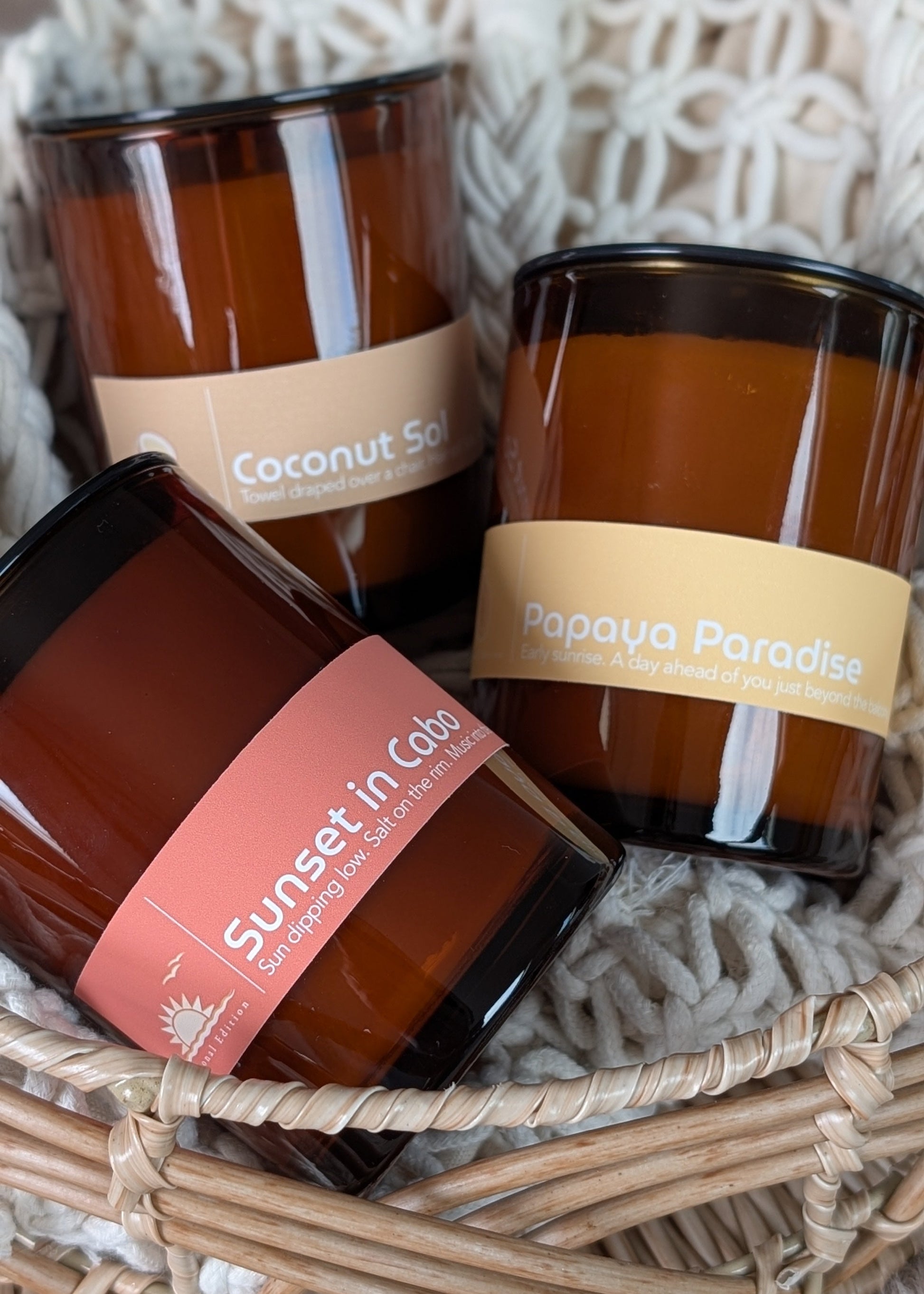Three summer scented amber glass candles with labels in a woven basket on a textured surface