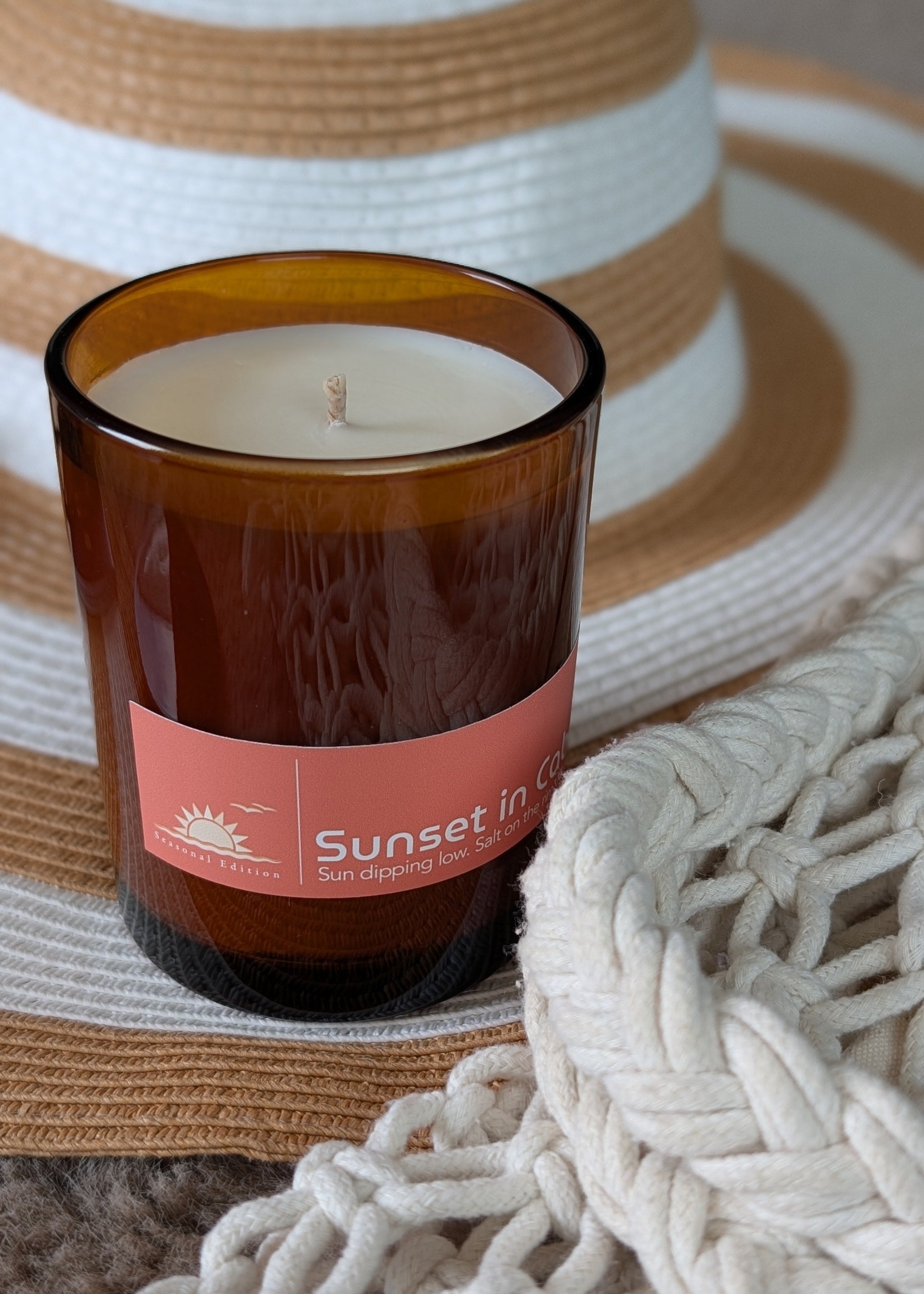 Candle in an amber glass vessel sitting on a beach hat and woven fabric