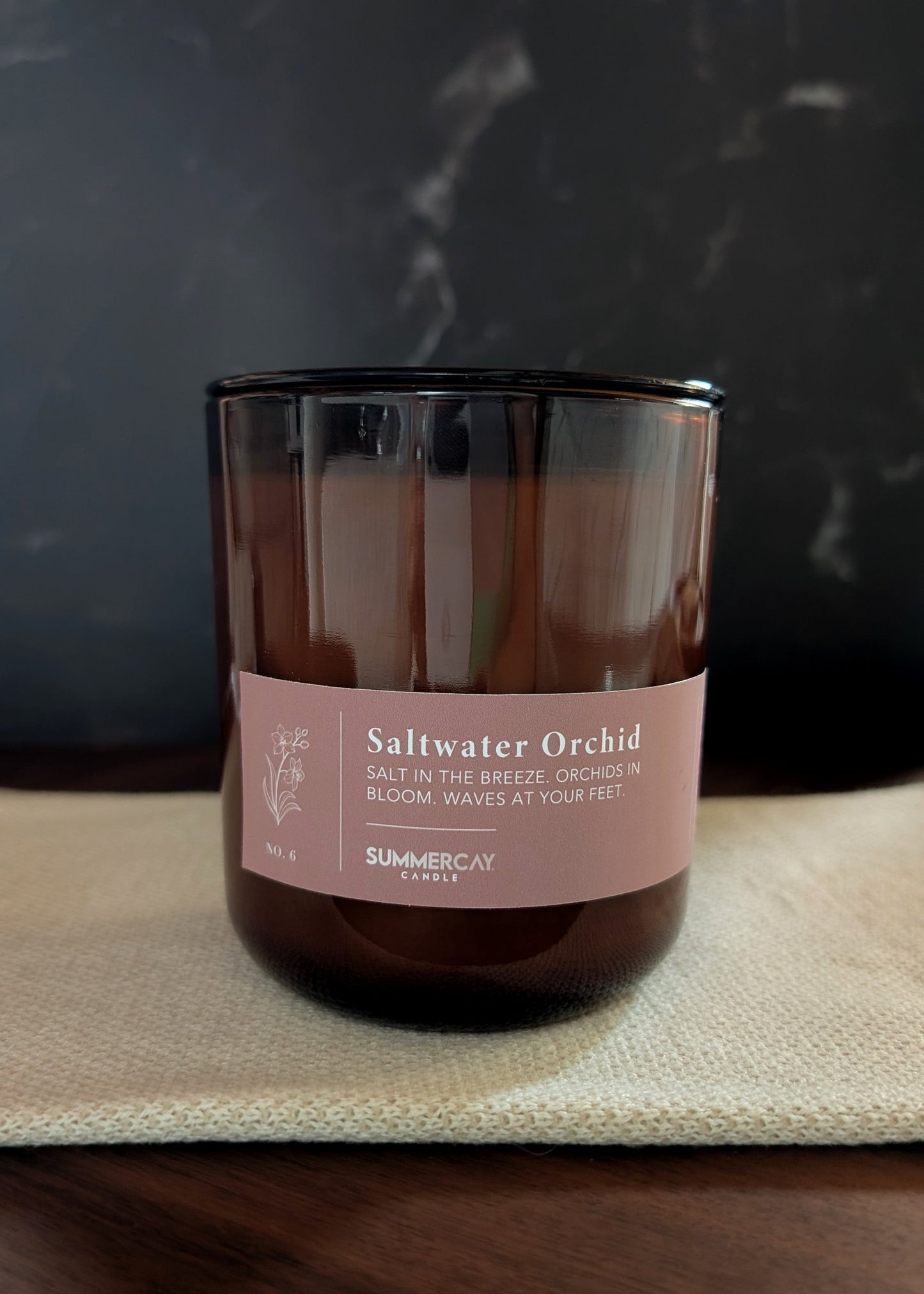 Amber glass candle labeled 'Saltwater Orchid' on a textured surface with a dark background