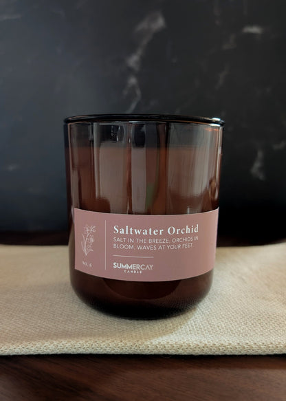Amber glass candle labeled 'Saltwater Orchid' on a textured surface with a dark background