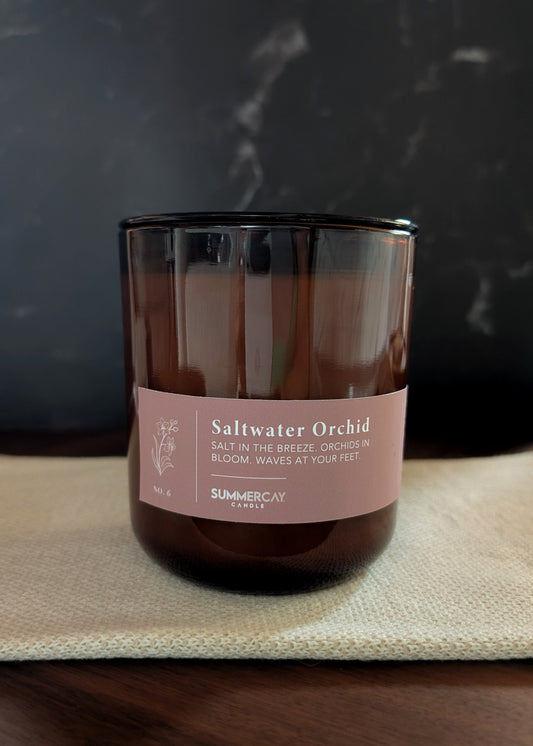 Amber glass candle labeled 'Saltwater Orchid' on a textured surface with a dark background