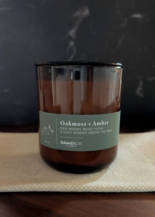 Candle labeled 'Oakmoss + Amber' on a textured surface with a dark background