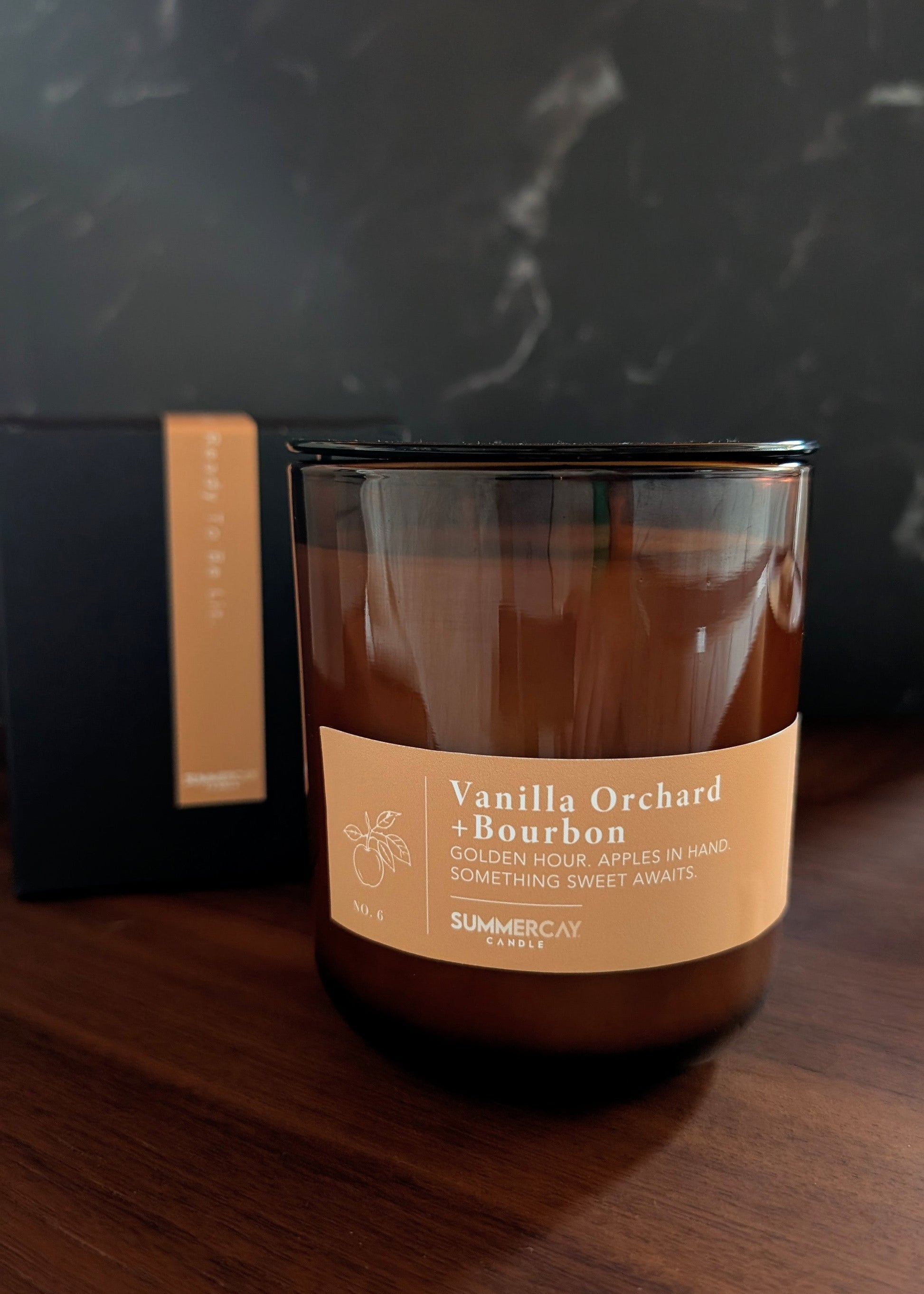 Vanilla Orchard and Bourbon candle in an amber vessel with he gift box in the back and a dark background.