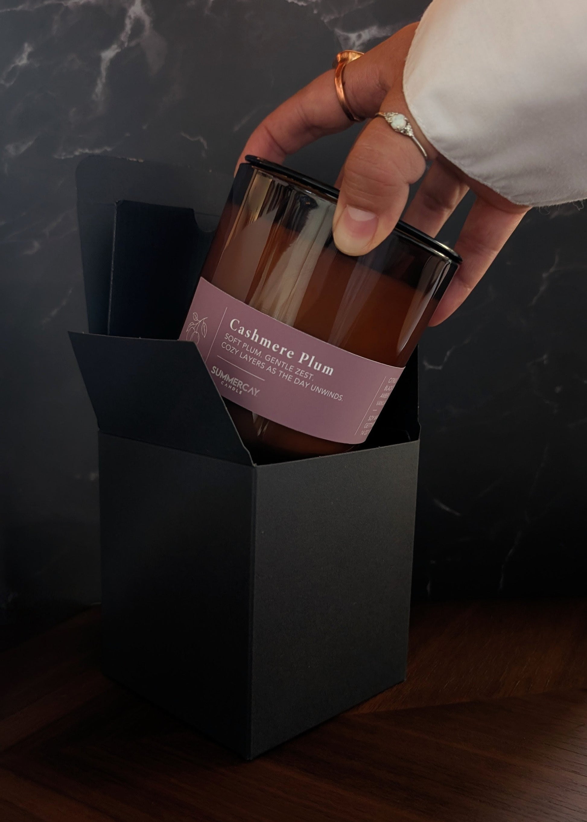 hand pulling out a Cashmere Plum candle from gift box with a dark background.