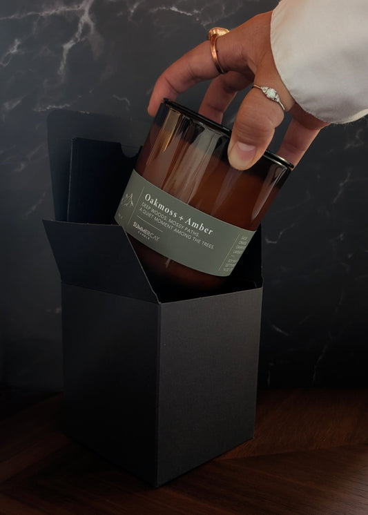 Hand pulling out amber glass candle named Oakmoss + Amber from black gift box.