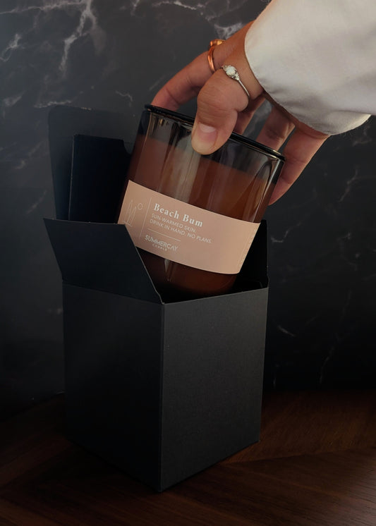 Hand pulling out an amber glass candle named Beach Bum out of a black gift box with a dark background.