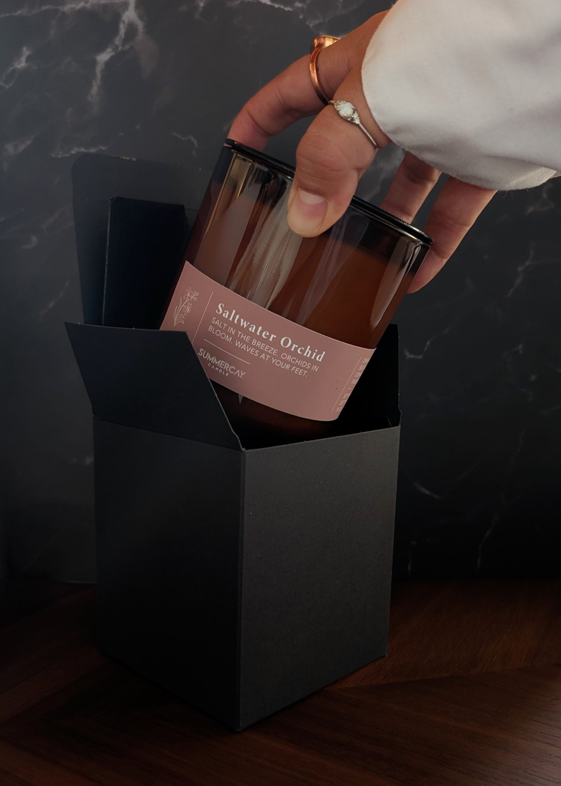 Hand holding a glass amber candle labeled 'Saltwater Orchid' with a black box on a dark surface.