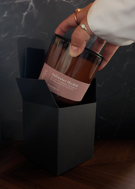 Hand holding a glass amber candle labeled 'Saltwater Orchid' with a black box on a dark surface.