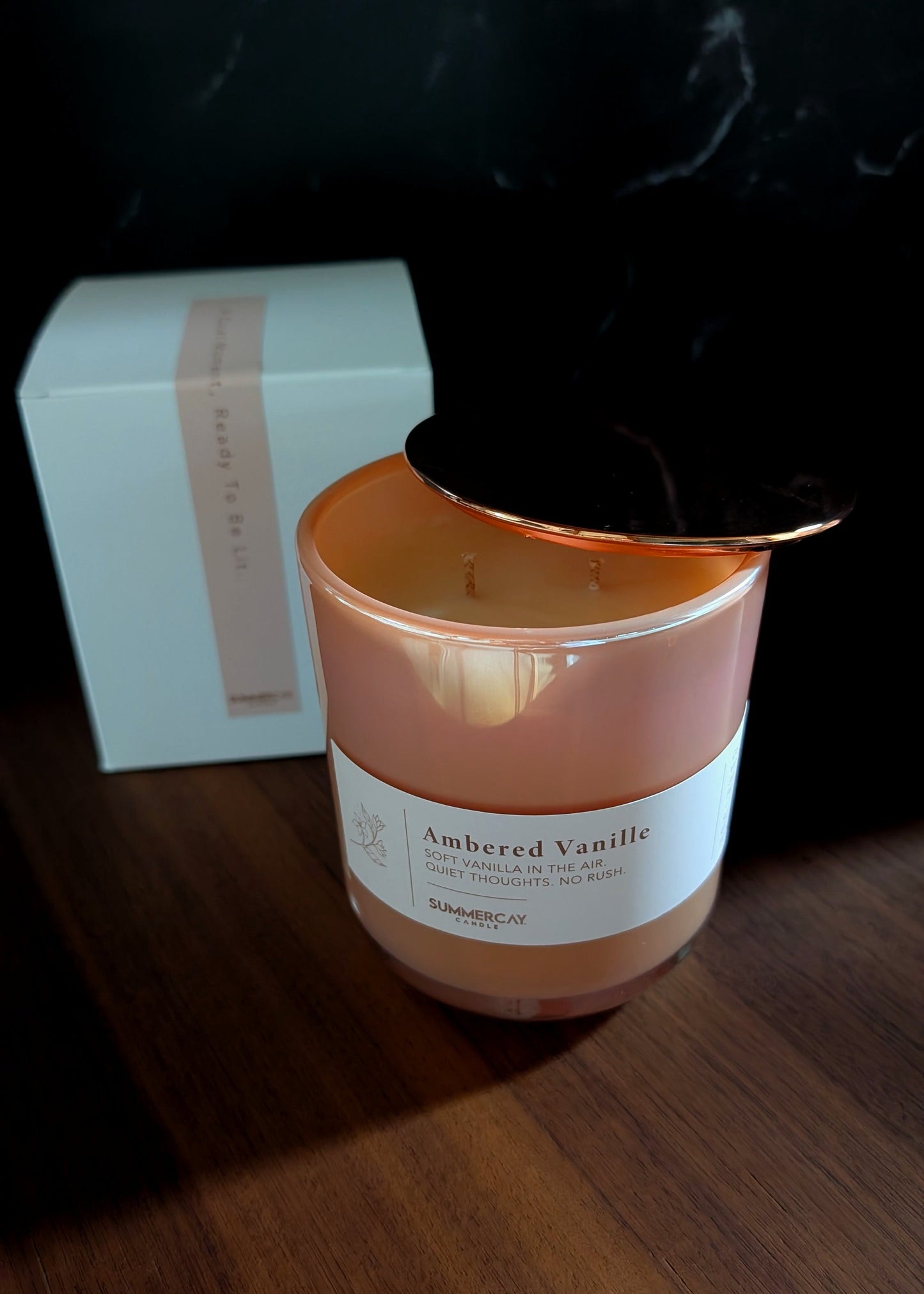 Candle labeled 'Ambered Vanille' with a white box in the background on a wooden surface.