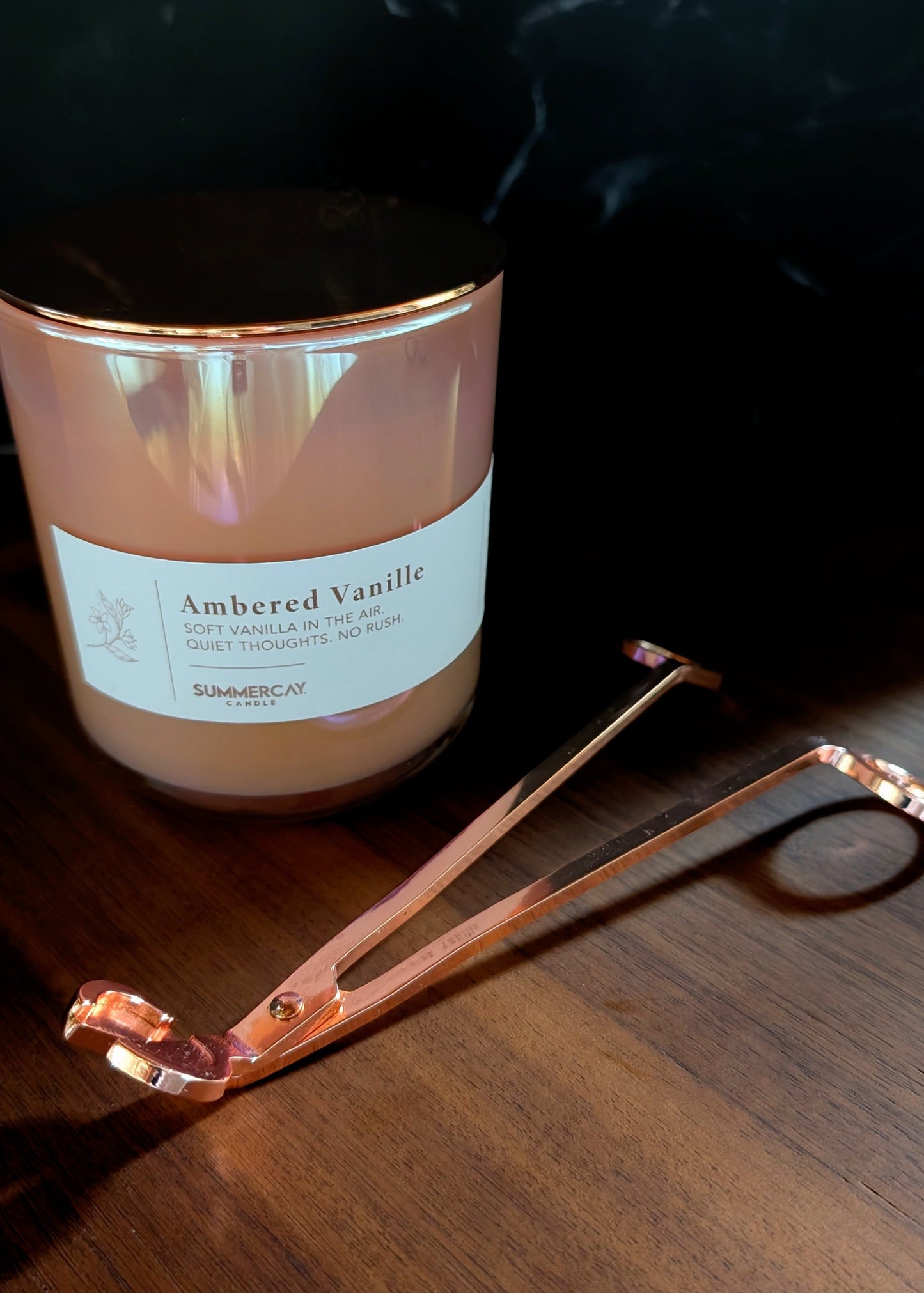 Rose gold wick trimmers next to a pink candle on a wooden surface and dark background.