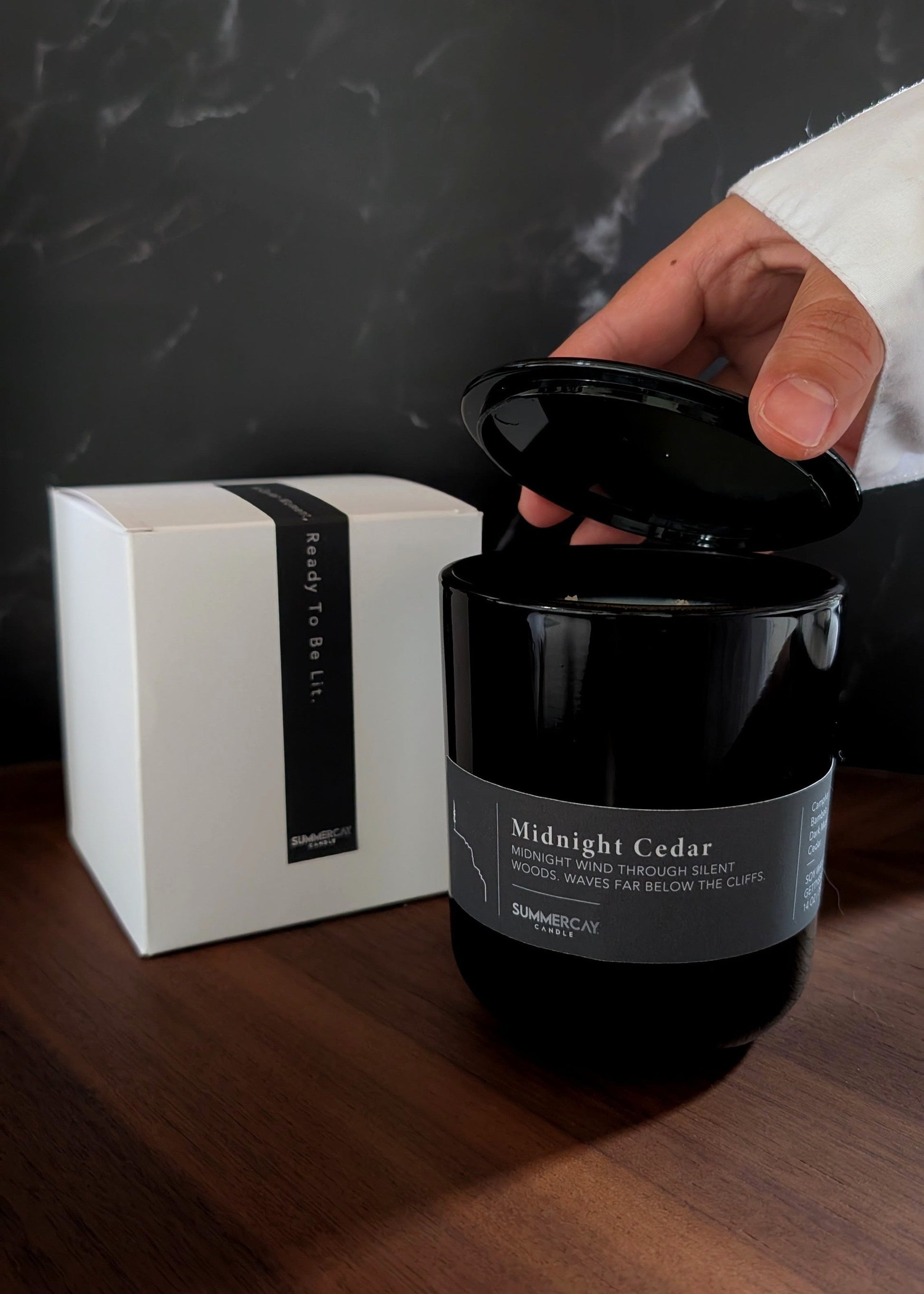 Black candle labeled 'Midnight Cedar' with a white box on a wooden surface.