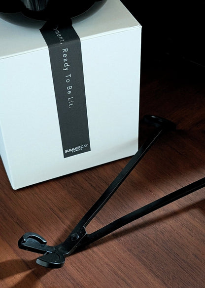 Black wick trimmers on a wooden surface next to a candle gift box and candle sitting on top.