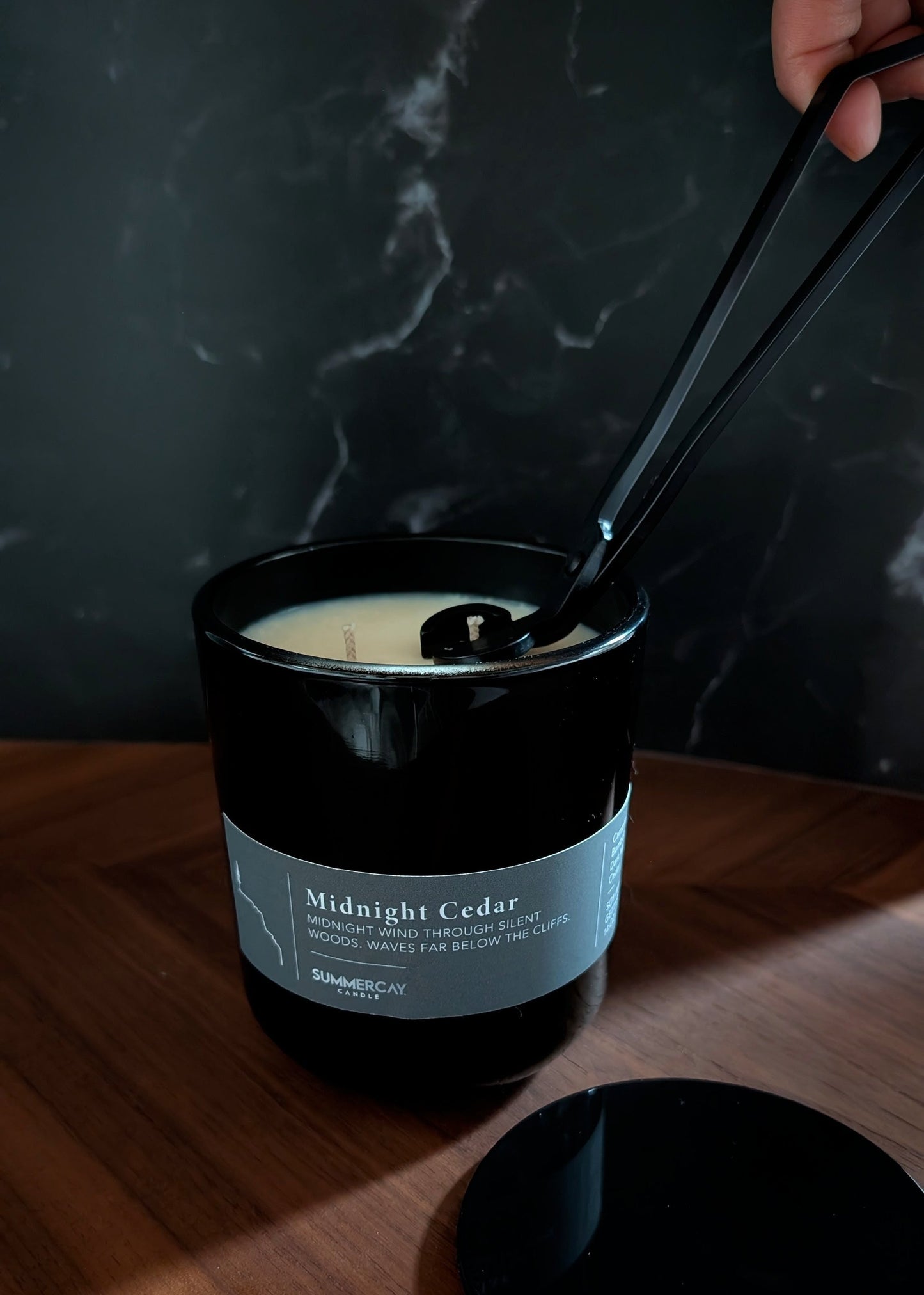 Candle labeled 'Midnight Cedar' with wick being trimmed by black wick trimmers on a wooden surface.