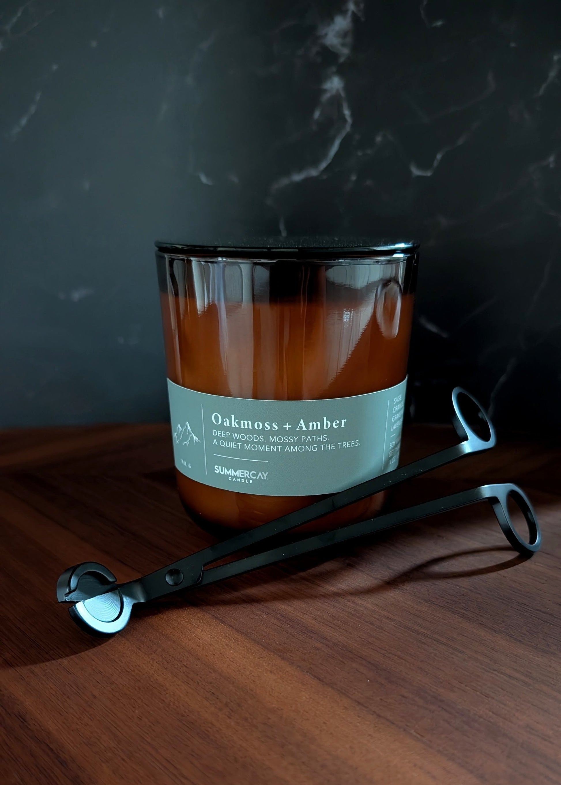 Candle labeled 'Oakmoss & Amber' on a wooden surface with a set of black wick trimmers with a dark background