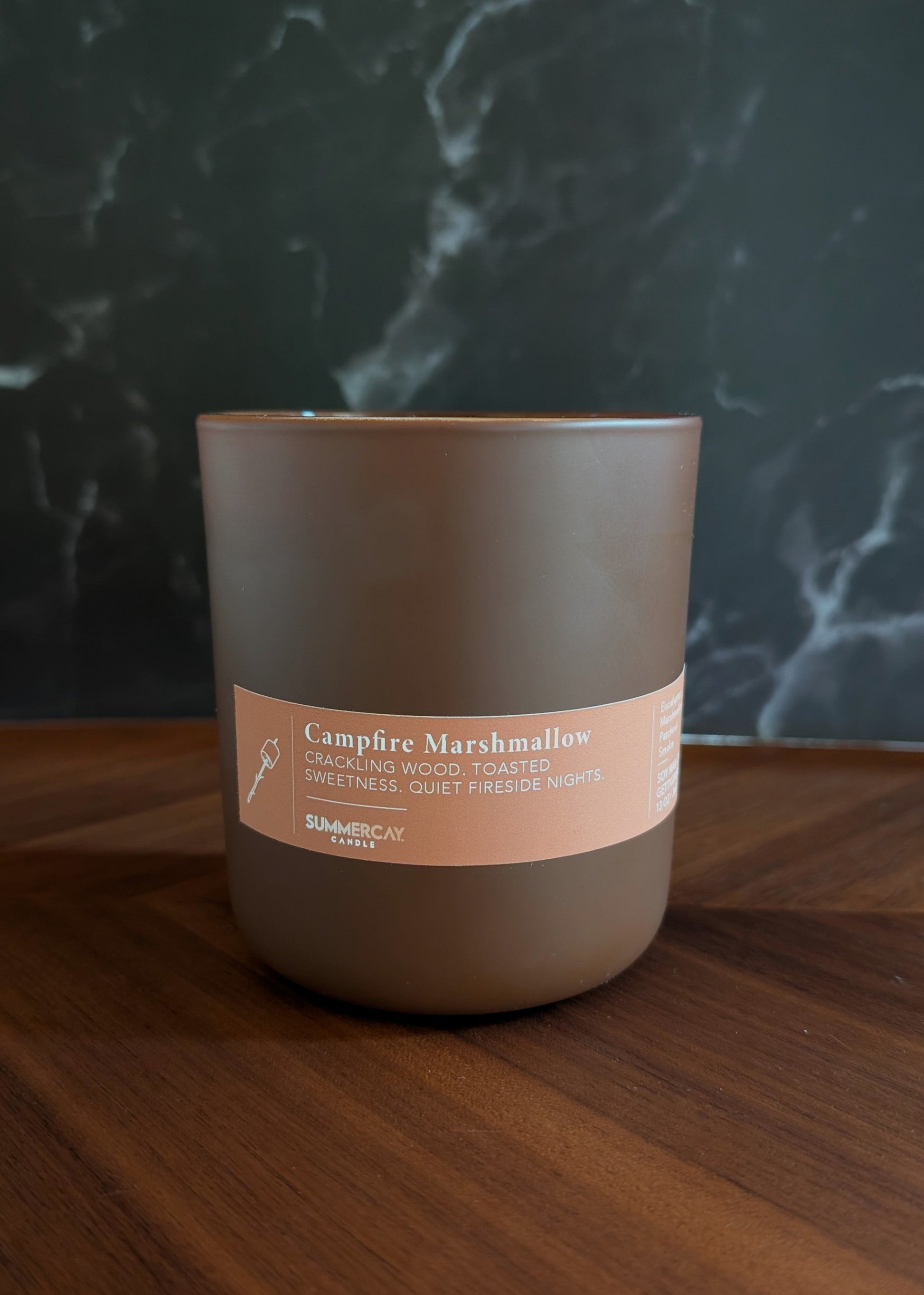 Candle labeled 'Campfire Marshmallow' on a wooden surface with a dark textured background