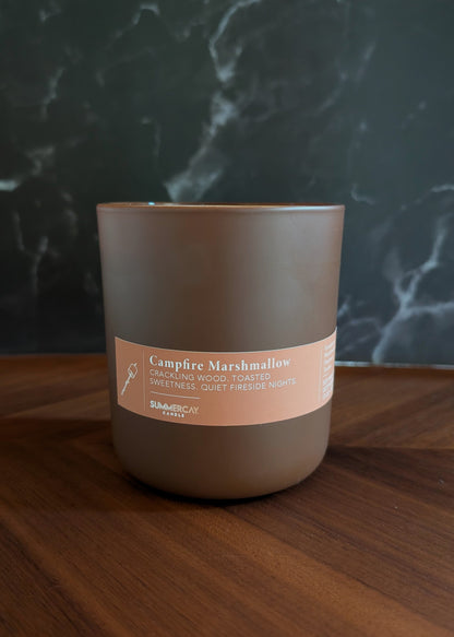 Candle labeled 'Campfire Marshmallow' on a wooden surface with a dark textured background