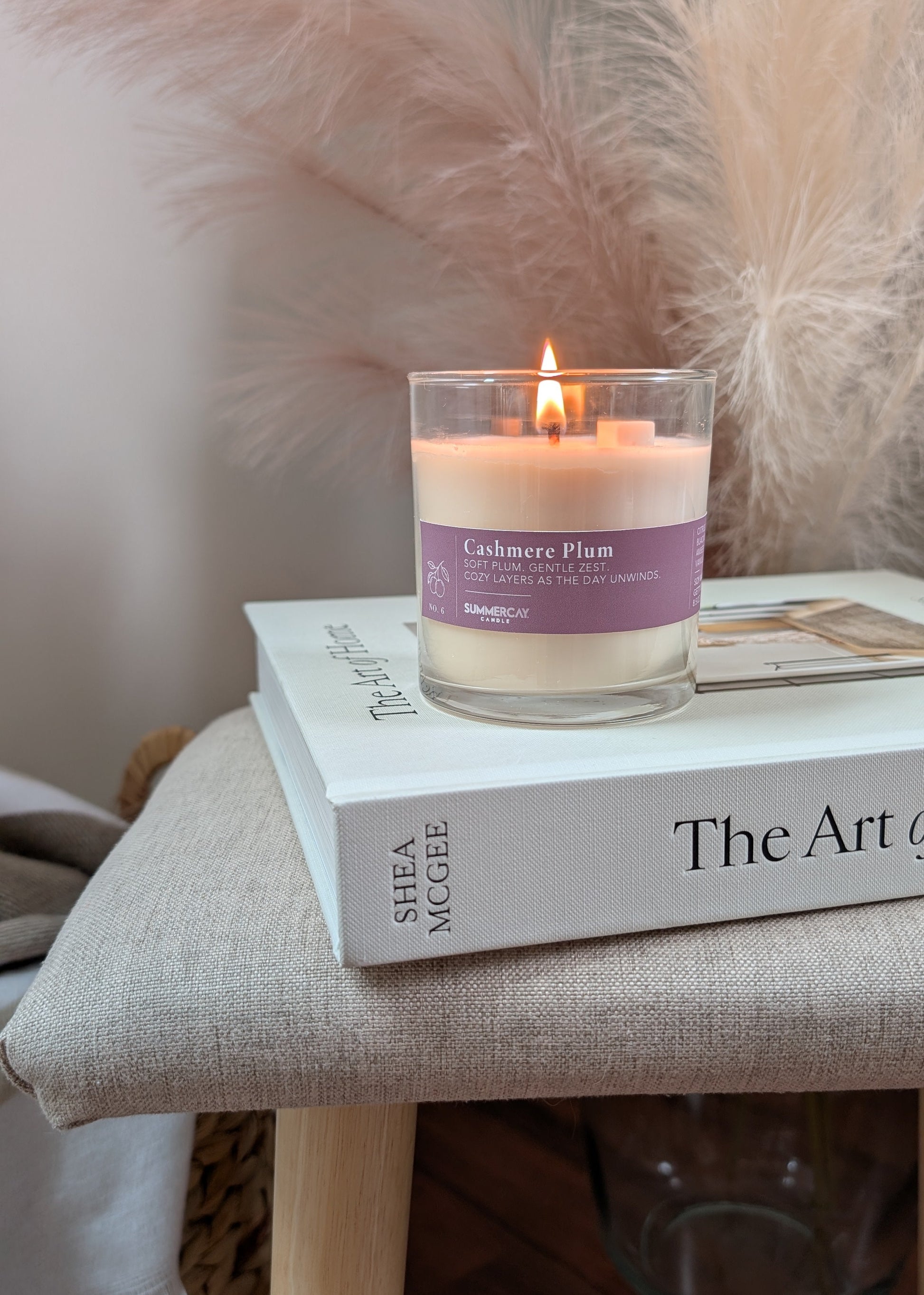A lit candle in a clear glass jar sits on top of a home decor book on a beige upholstered stool. The candle label reads 'Cashmere Plum' and shows the brand SummerCay Candle.
