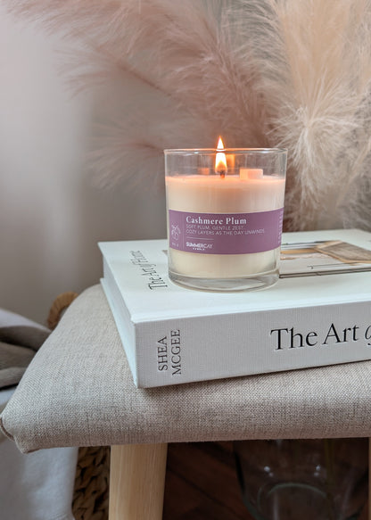 A lit candle in a clear glass jar sits on top of a home decor book on a beige upholstered stool. The candle label reads 'Cashmere Plum' and shows the brand SummerCay Candle.