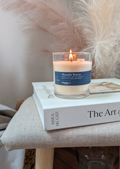 A lit candle in a clear glass jar sits on top of a book on a beige upholstered stool. The candle label reads 'Moonlit Waves'.