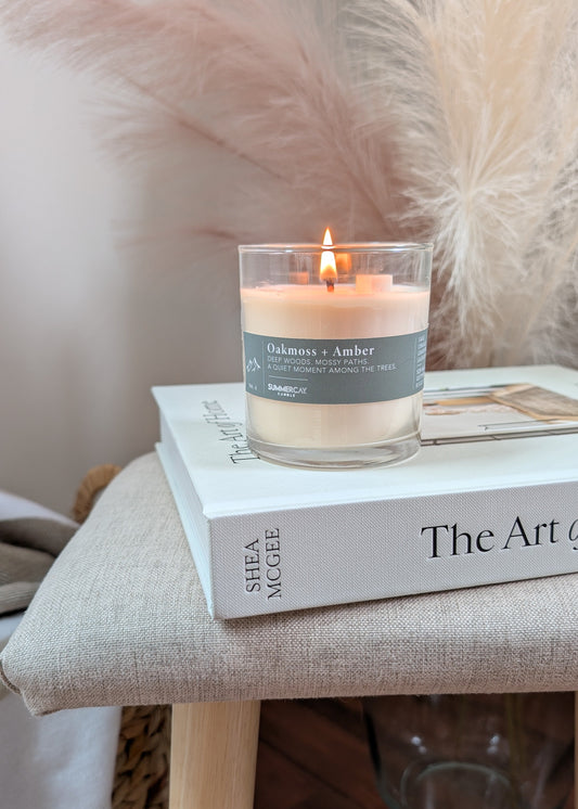 A lit candle in a clear glass jar sits on top of a white book on a beige upholstered stool. The candle label reads 'Oakmoss + Amber' and the book spine shows 'SHEA MCGEE'.
