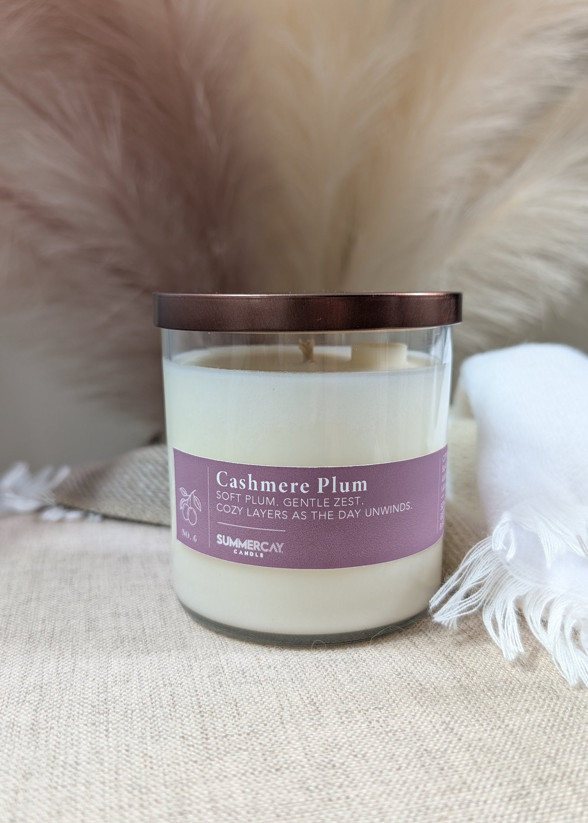 A candle in a clear glass jar with a bronze lid and a deep plum colored label reading 'Cashmere Plum' on a beige fabric surface with soft neutral props in the background.