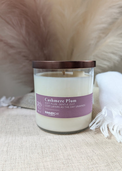 A candle in a clear glass jar with a bronze lid and a deep plum colored label reading 'Cashmere Plum' on a beige fabric surface with soft neutral props in the background.